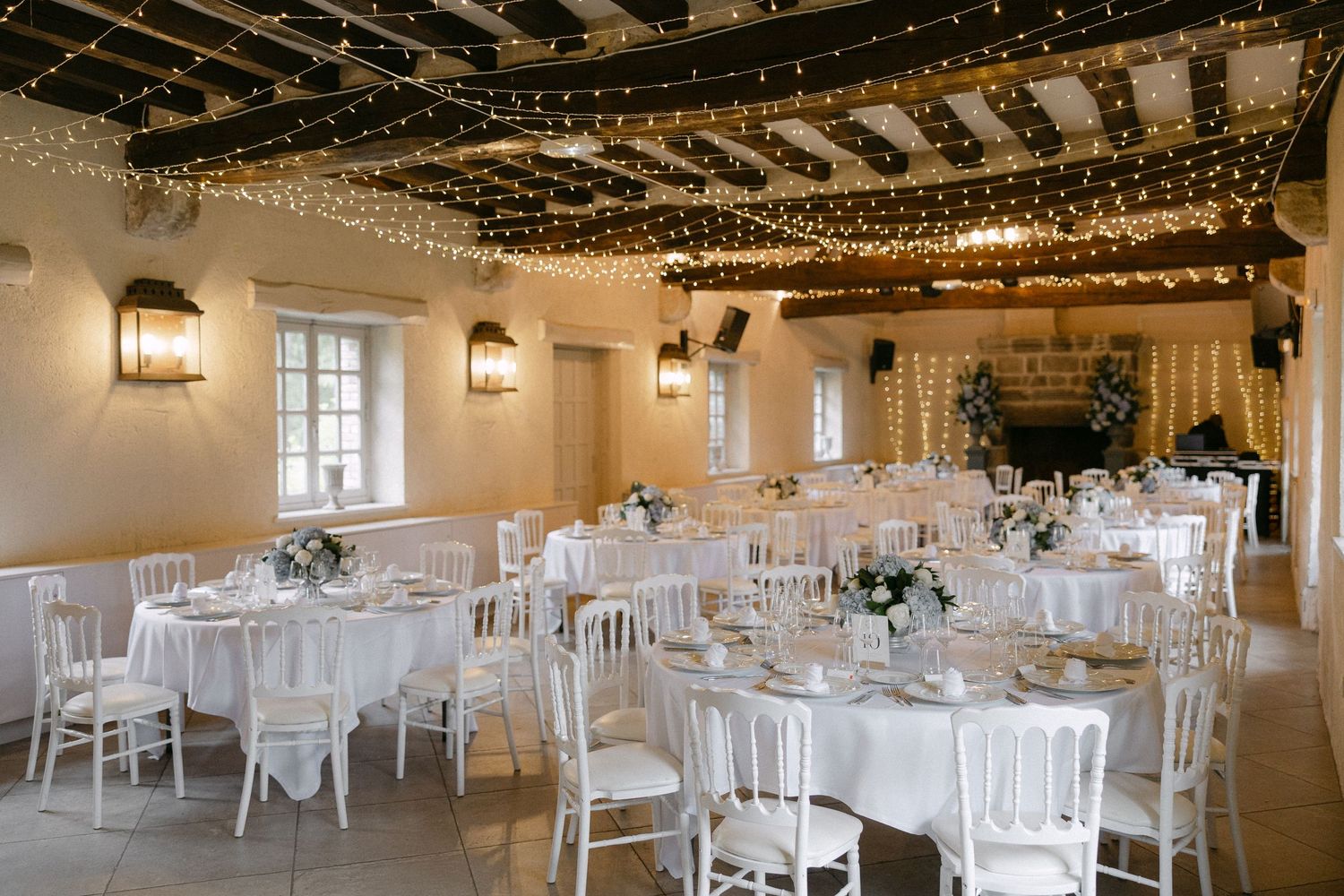 Romantic wedding reception hall with fairy lights on beamed ceiling, white round tables and chiavari chairs.