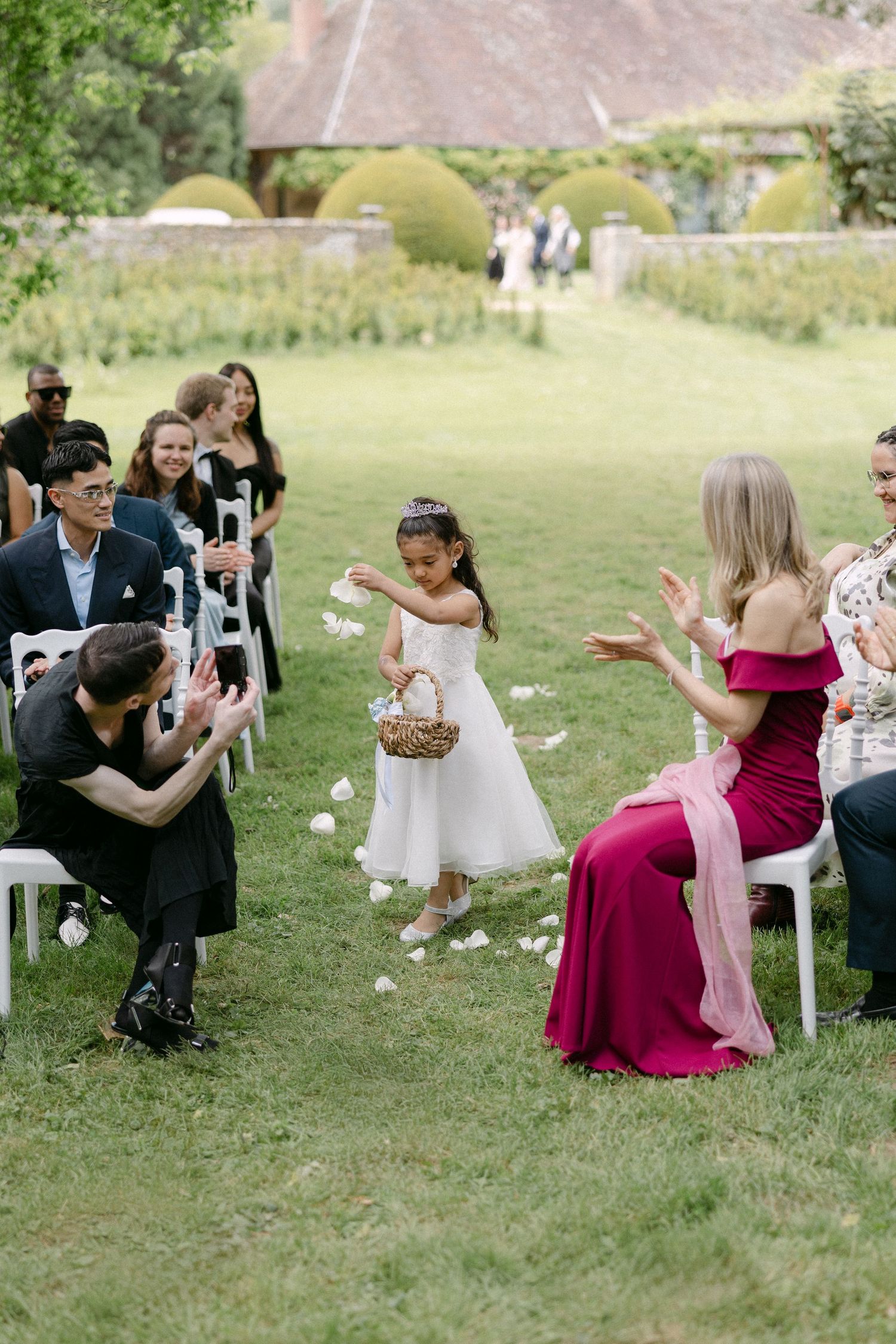 Adorable flower girl tossing white petals down the aisle at an outdoor garden wedding ceremony.