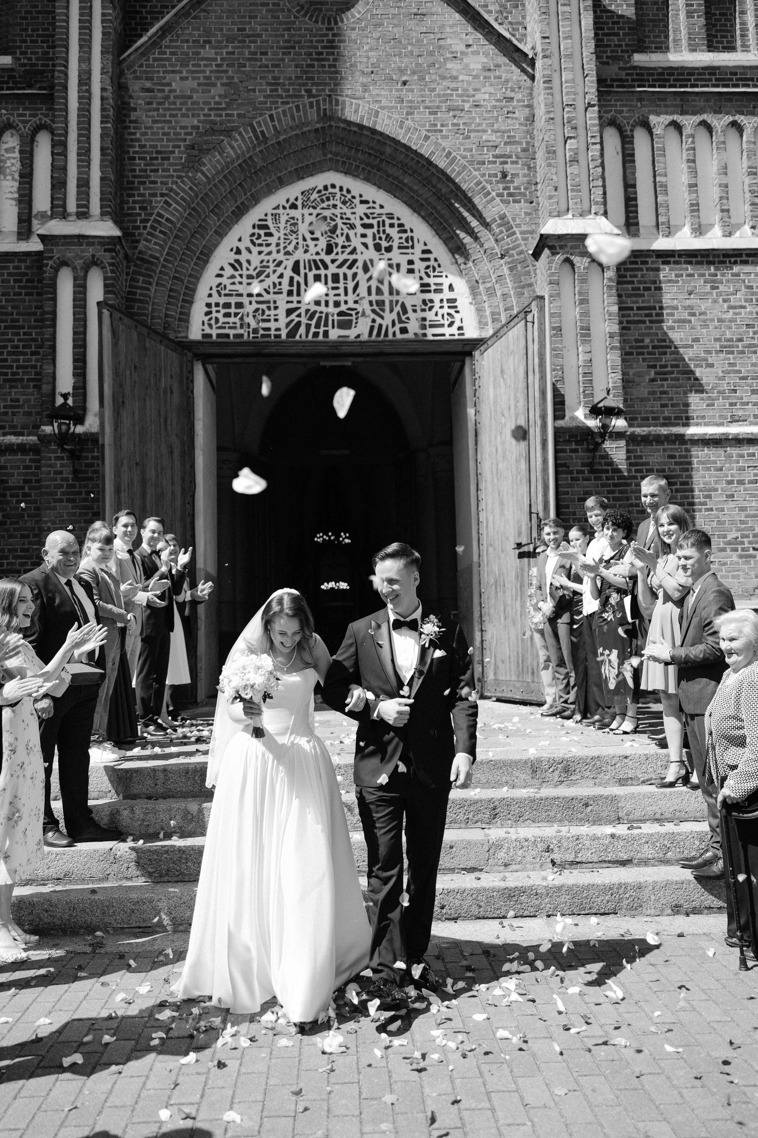 Bride and groom exit brick church as guests toss flower petals in black and white wedding photo.