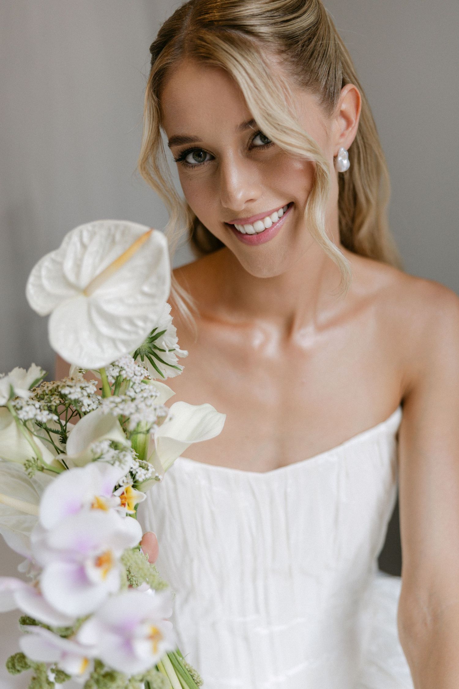 Smiling bride holding a white anthurium and orchid bouquet in a strapless wedding gown with pearl earrings.