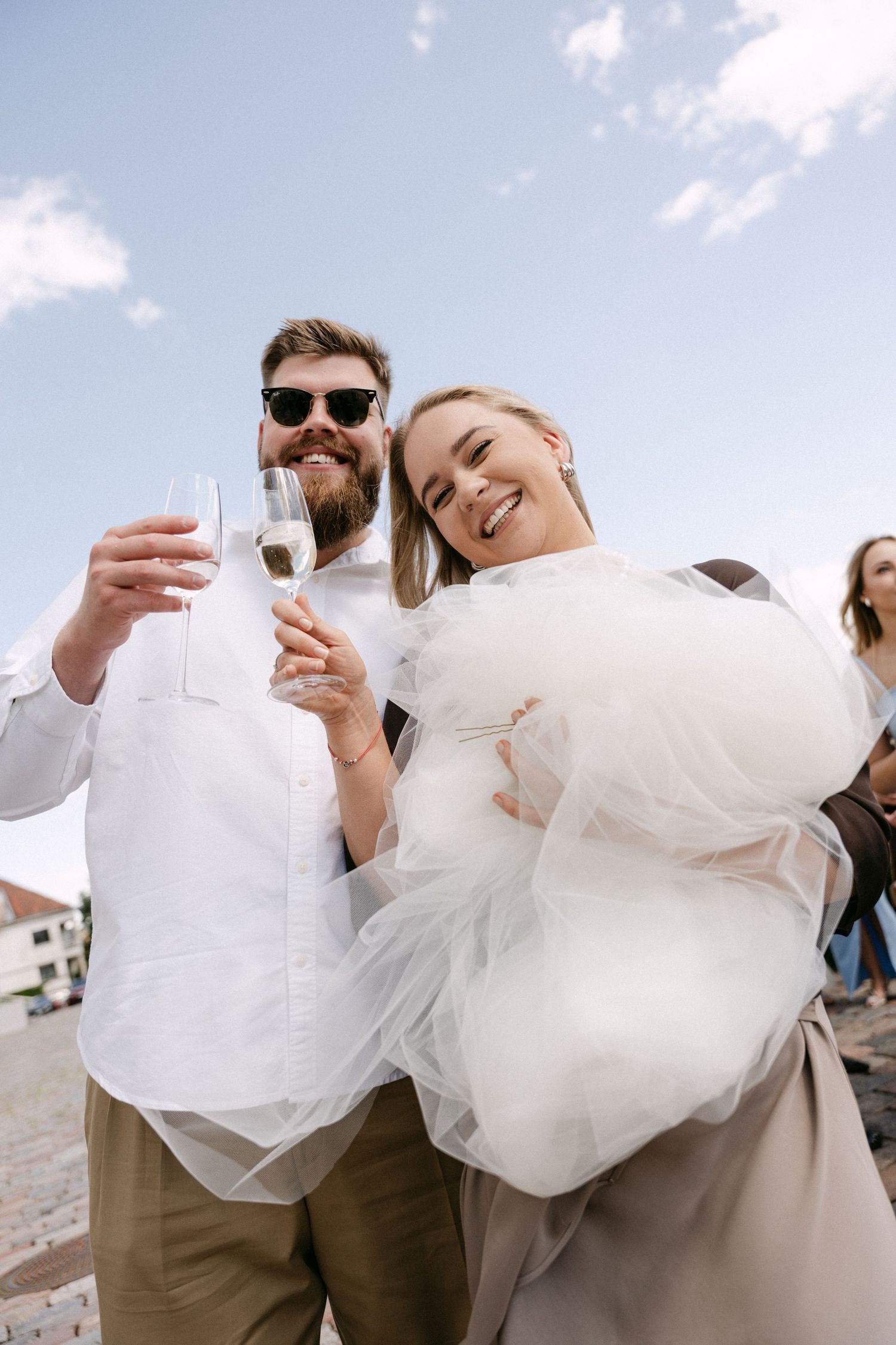 Smiling bride and bearded groom in sunglasses toasting with champagne glasses outdoors under blue sky.