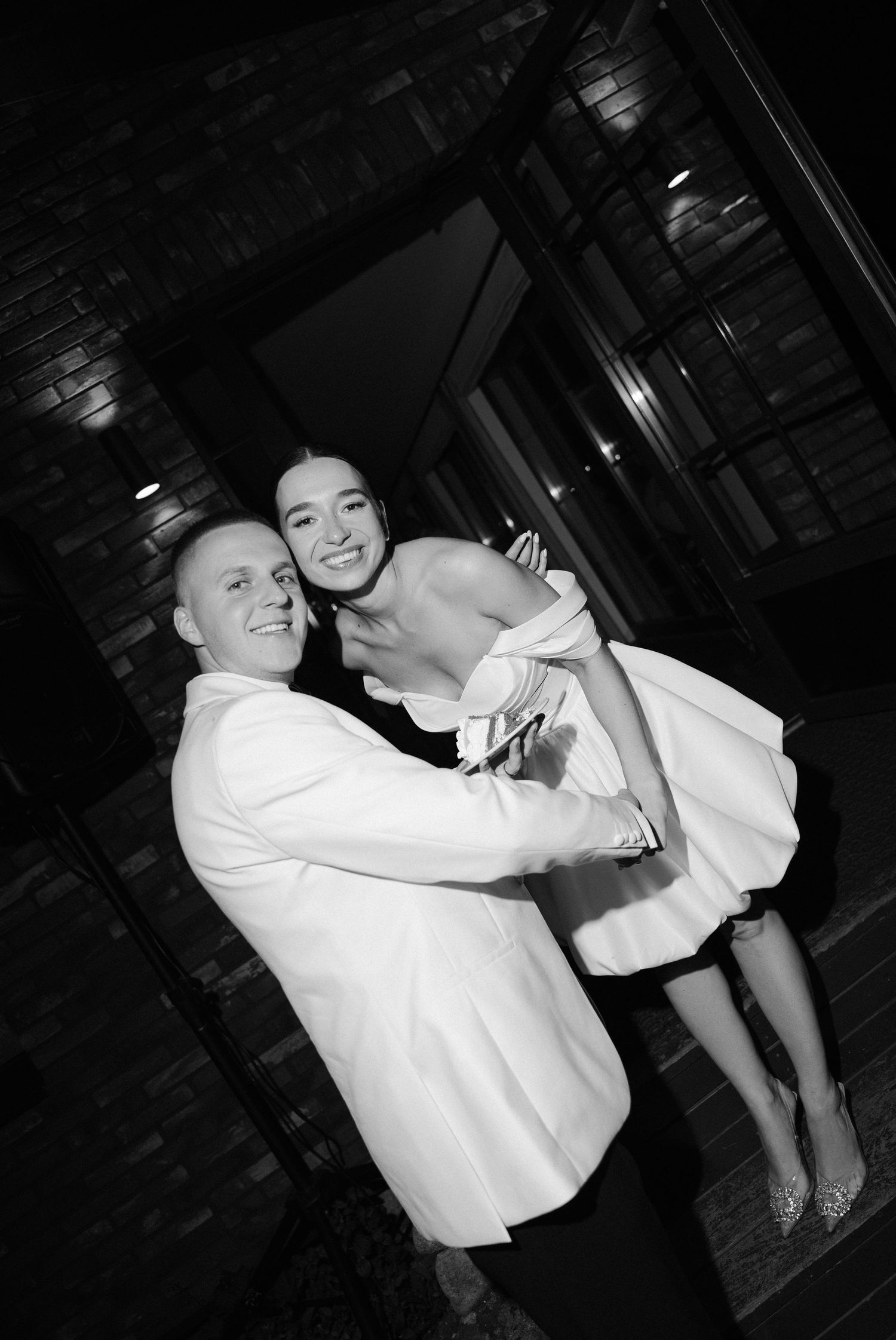 Black and white photo of groom lifting smiling bride in short white dress outside venue at night.