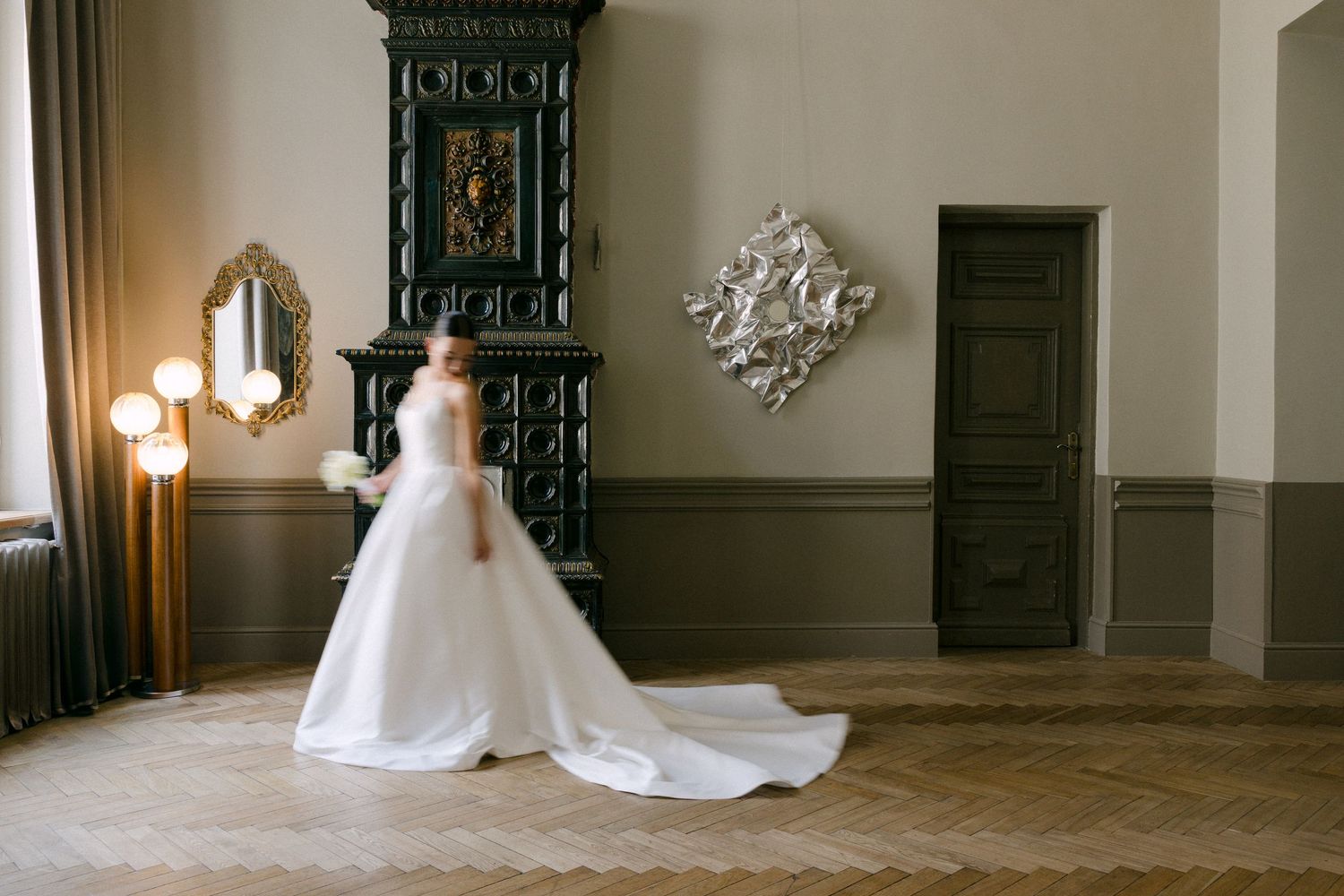 Bride in elegant white ball gown with long train standing in ornate room with decorative fireplace and mirror.