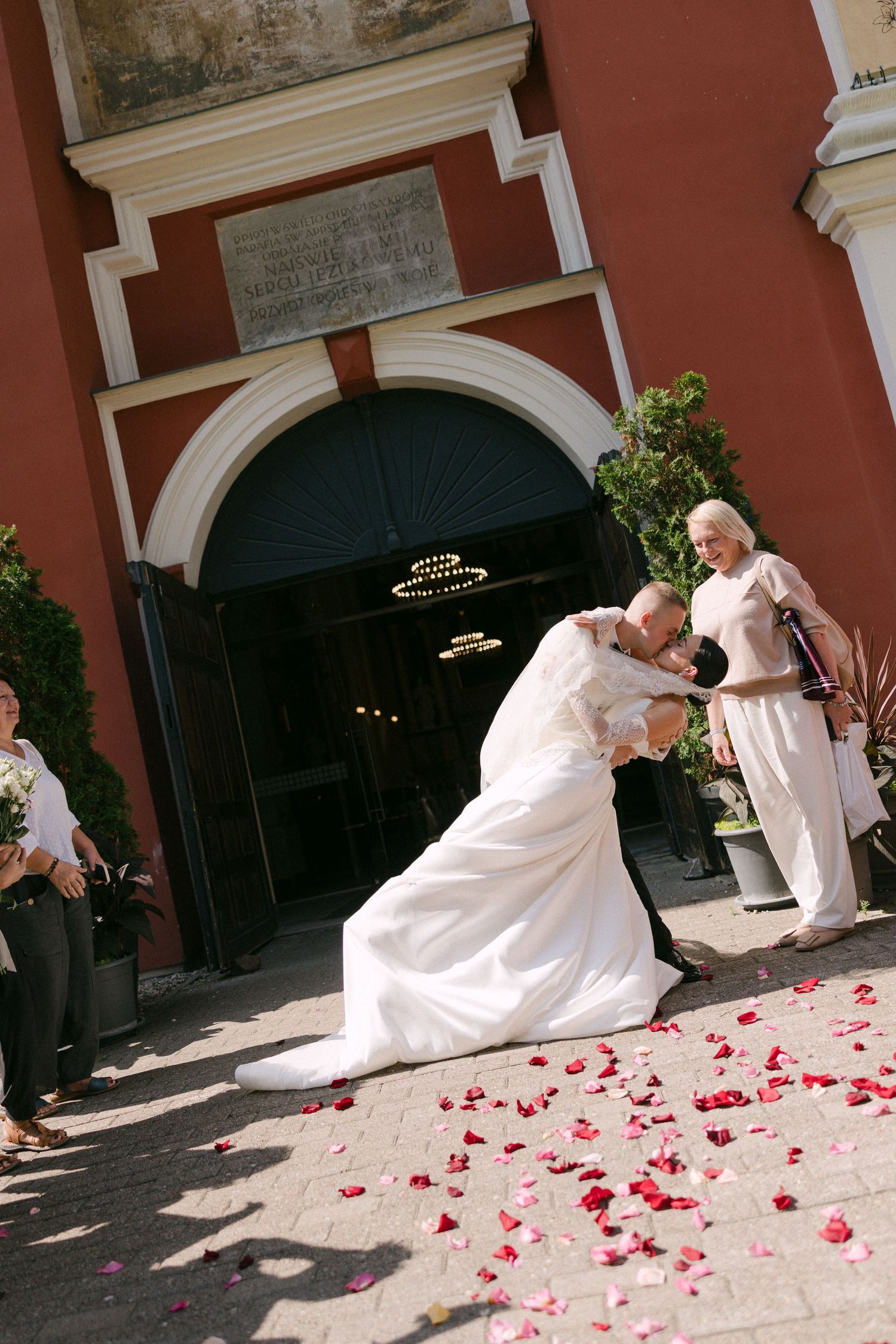Groom dips bride for romantic kiss outside church entrance scattered with red rose petals.