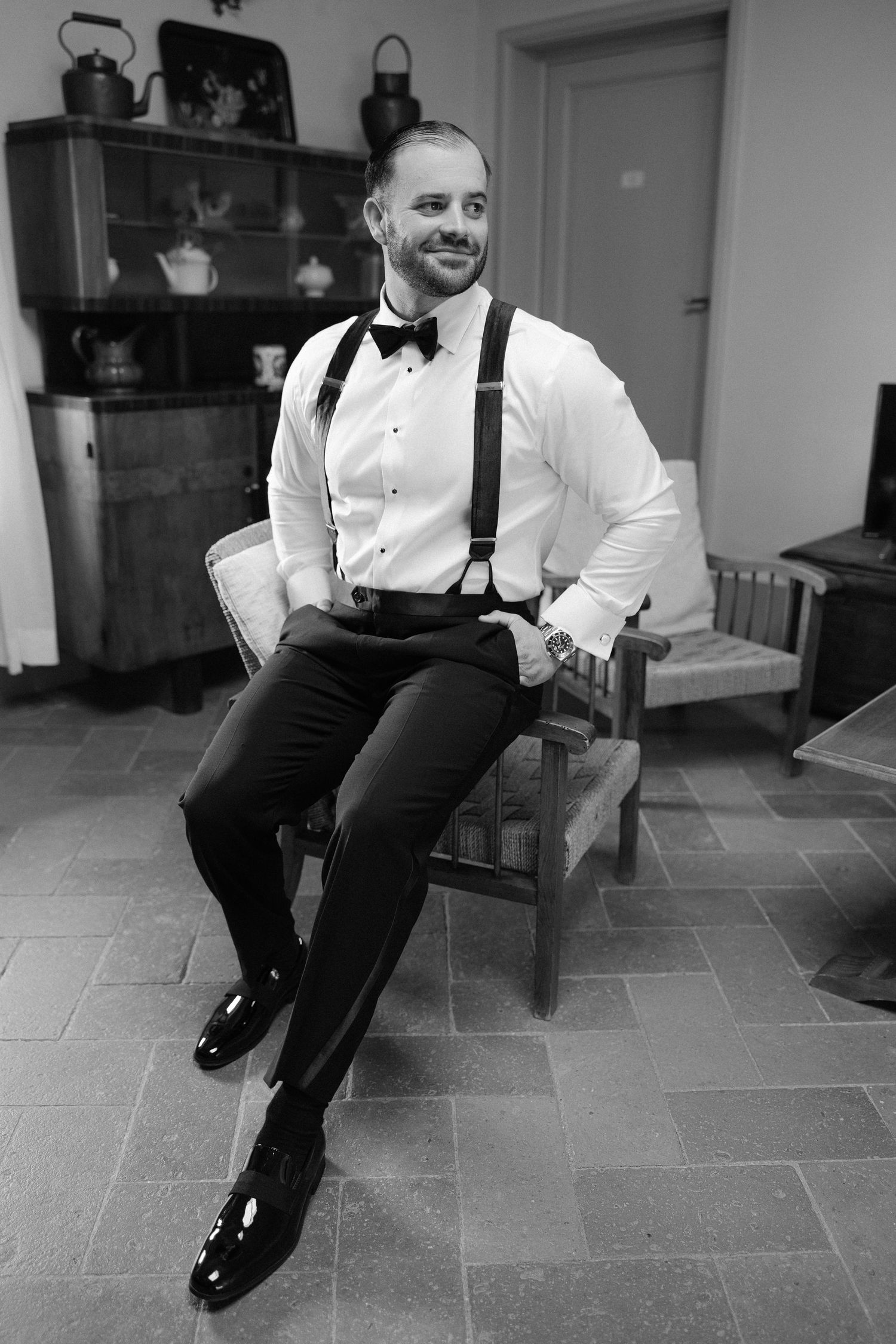 Dapper groom in black tuxedo with suspenders and bow tie sitting in chair smiling during wedding preparations.