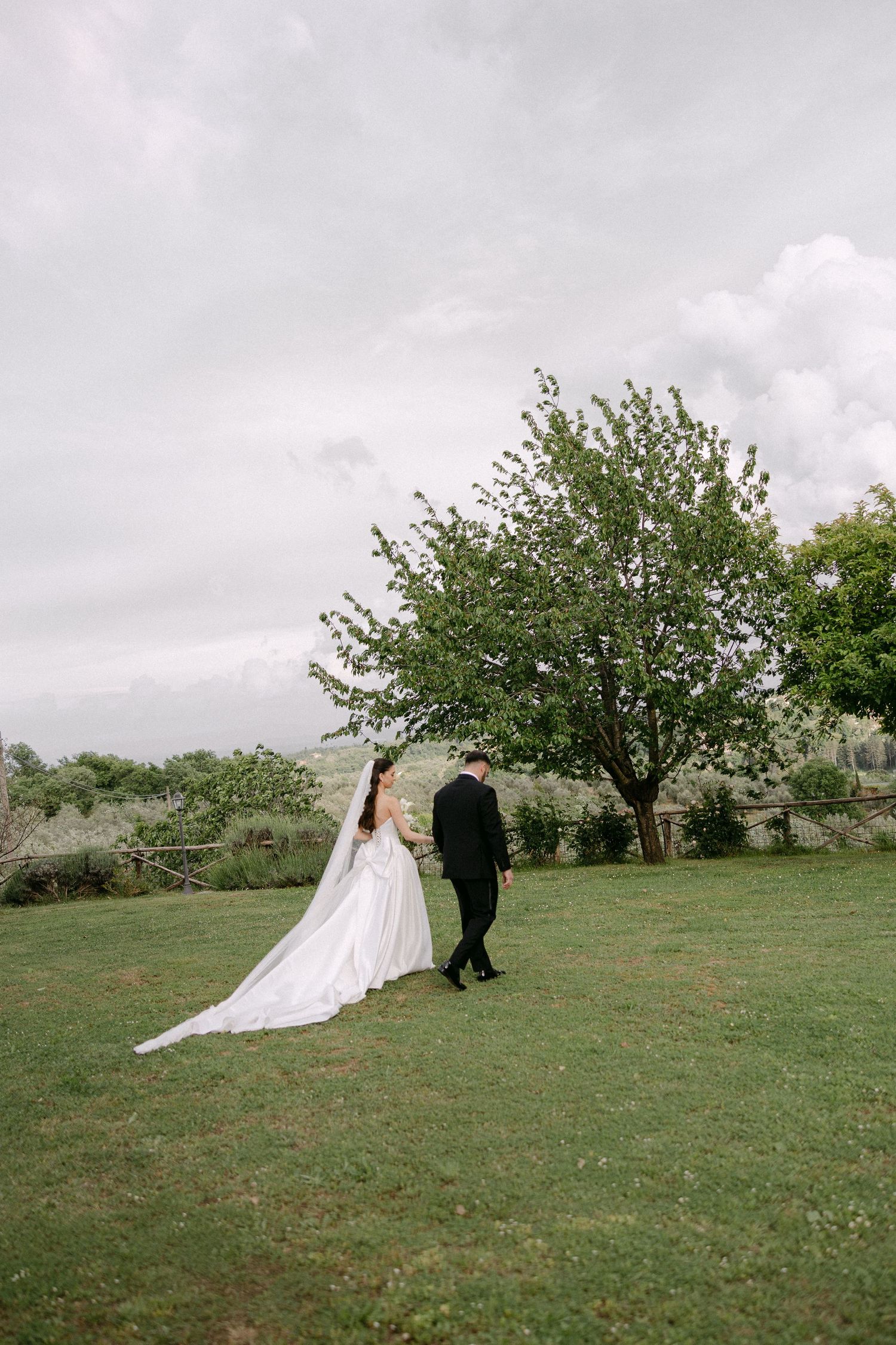 Bride and groom walking hand in hand across green lawn with dramatic veil train flowing behind.