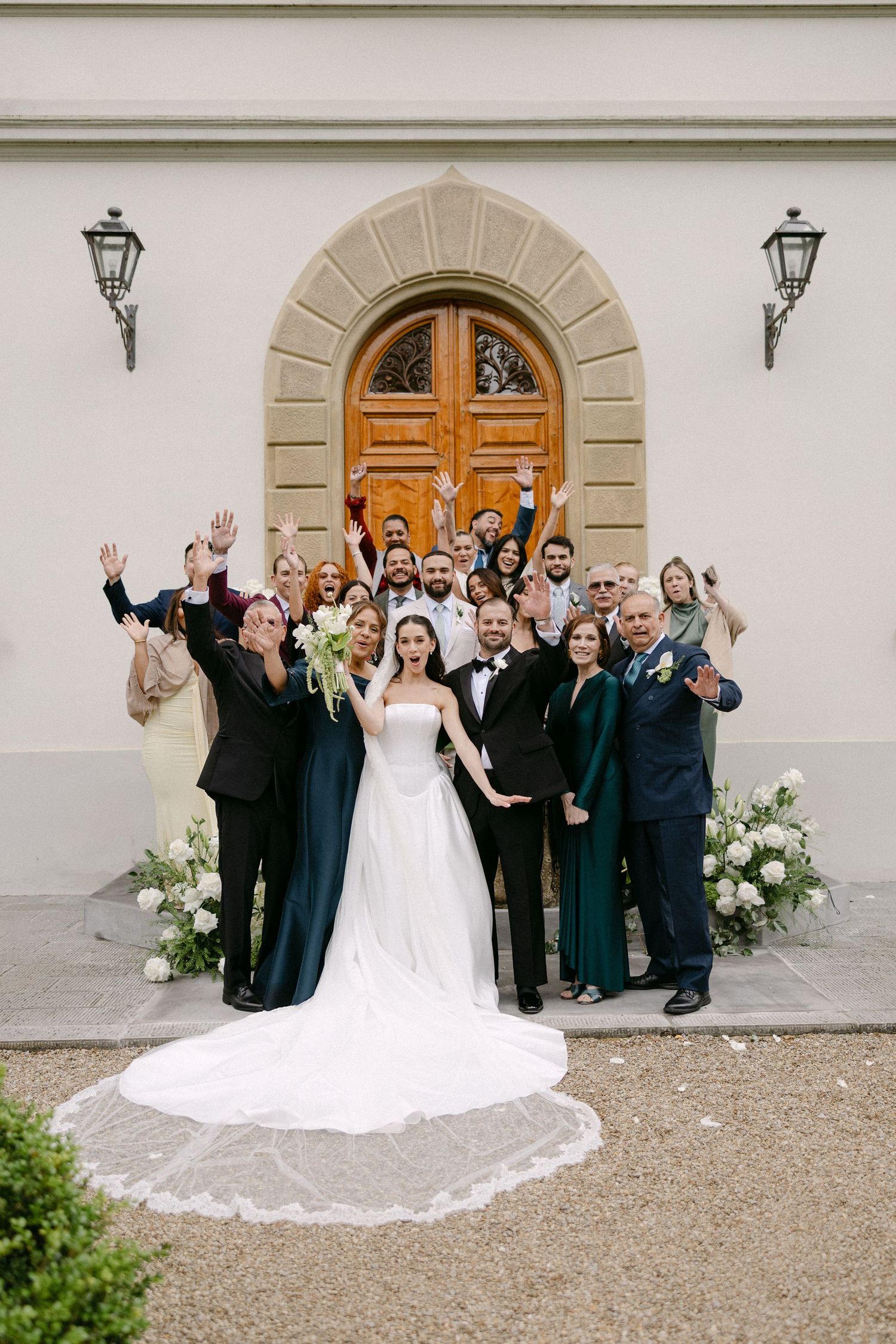 Wedding party celebrating with raised hands in front of elegant arched doorway with white floral arrangements.