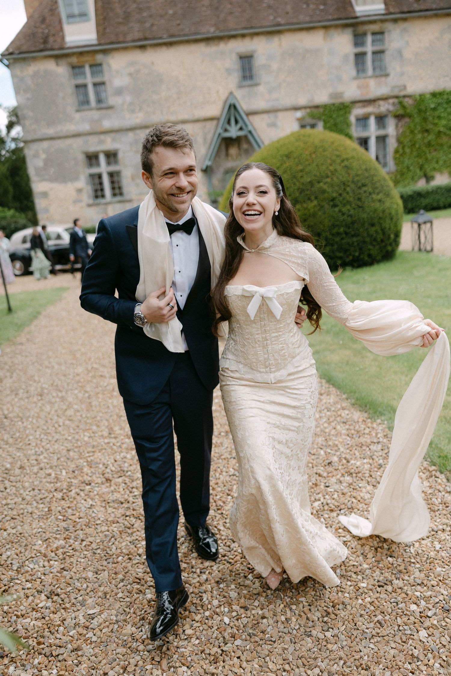 Smiling bride and groom walking together outside a stone manor house, bride wearing elegant lace wedding gown.