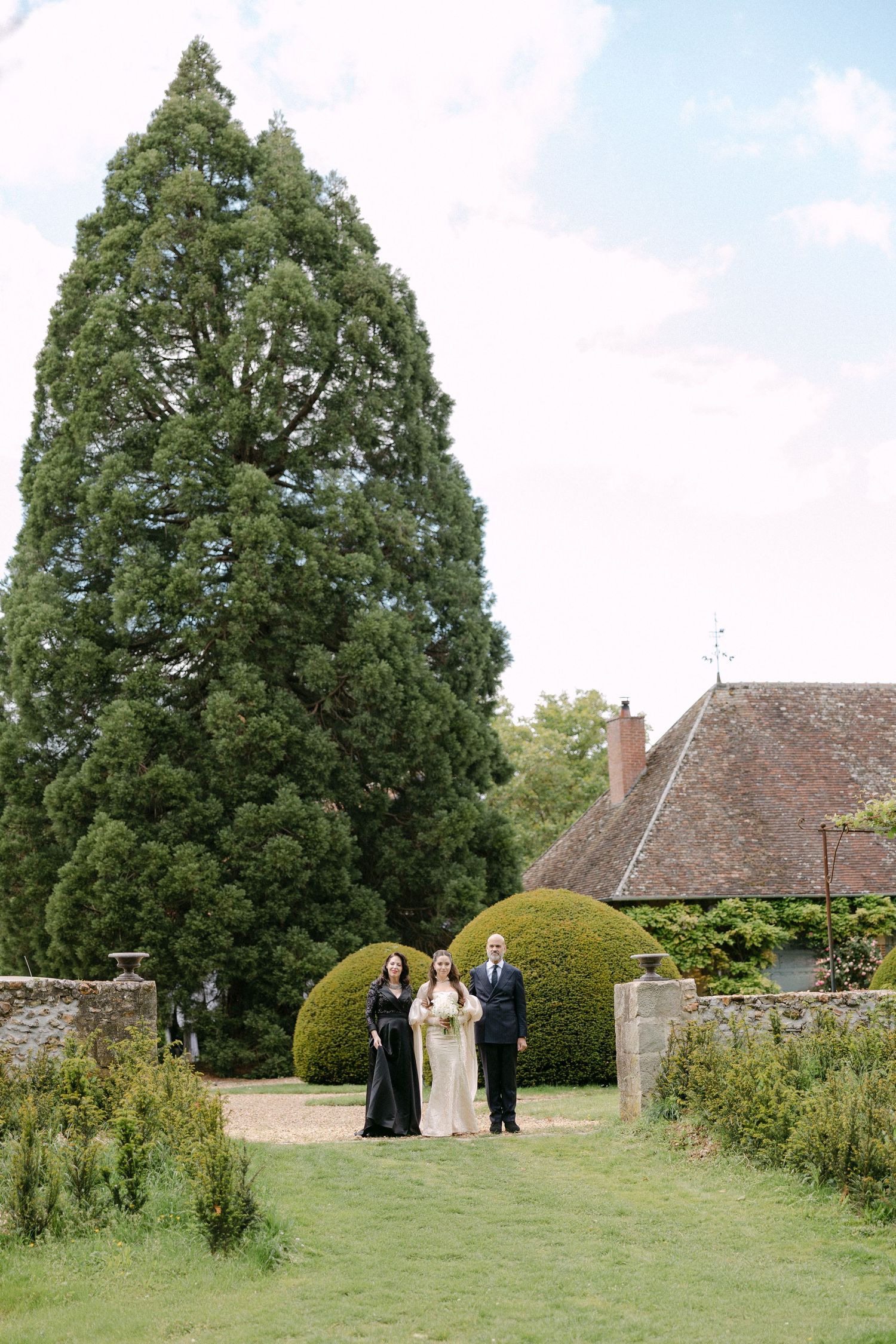 Bride in white gown walks with two guests through lush English country estate gardens.