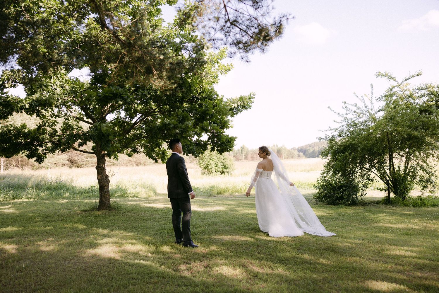 Bride in flowing white gown and groom in black suit walking together under trees in green garden.