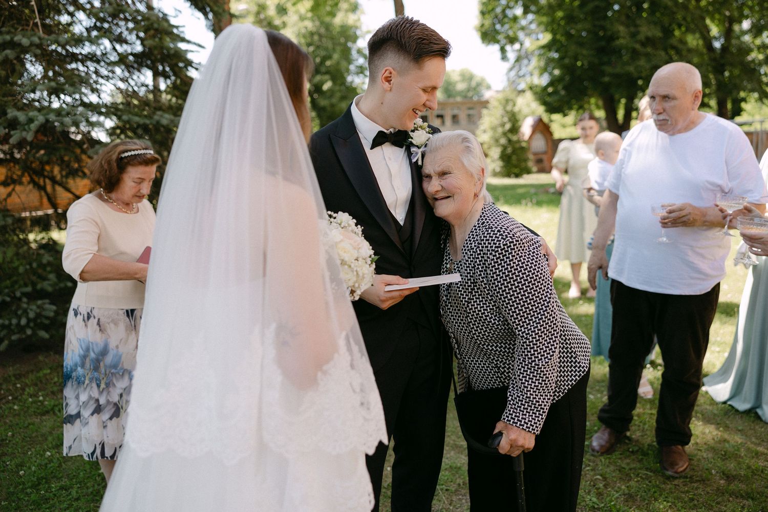Bride and groom greeting elderly woman guest at outdoor wedding reception on green lawn.
