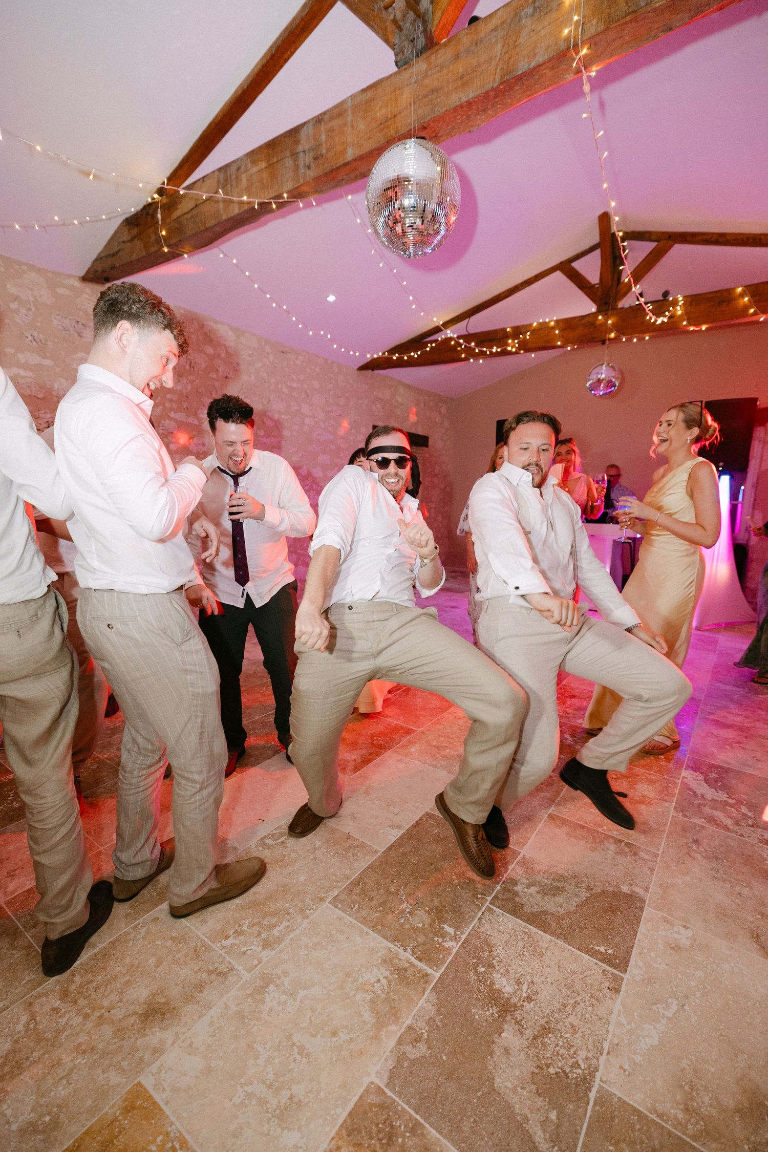 Groomsmen in beige suits dancing energetically at wedding reception under disco ball and fairy lights.