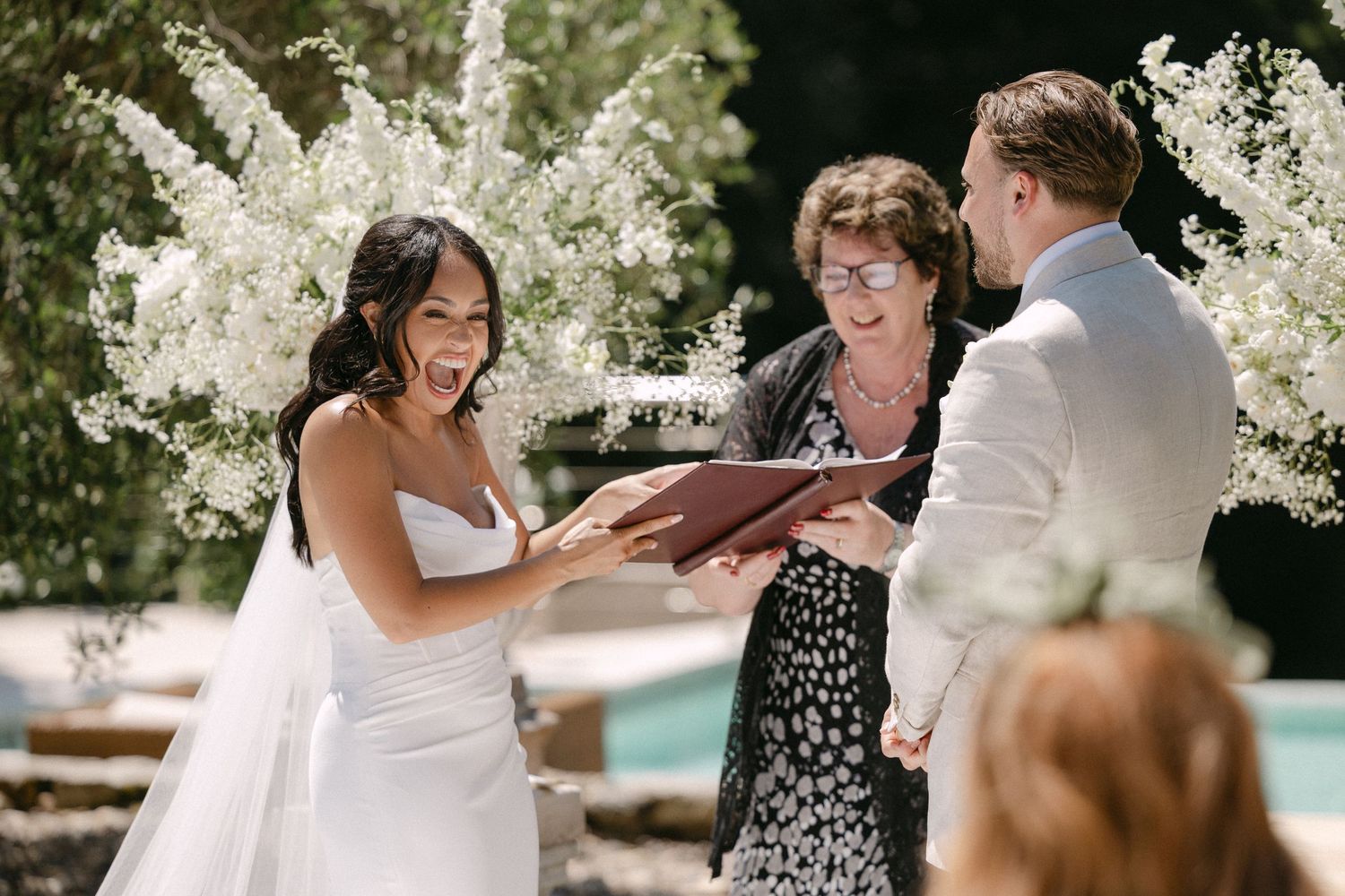 Bride and groom exchanging vows with officiant at outdoor wedding ceremony surrounded by white floral arrangements.