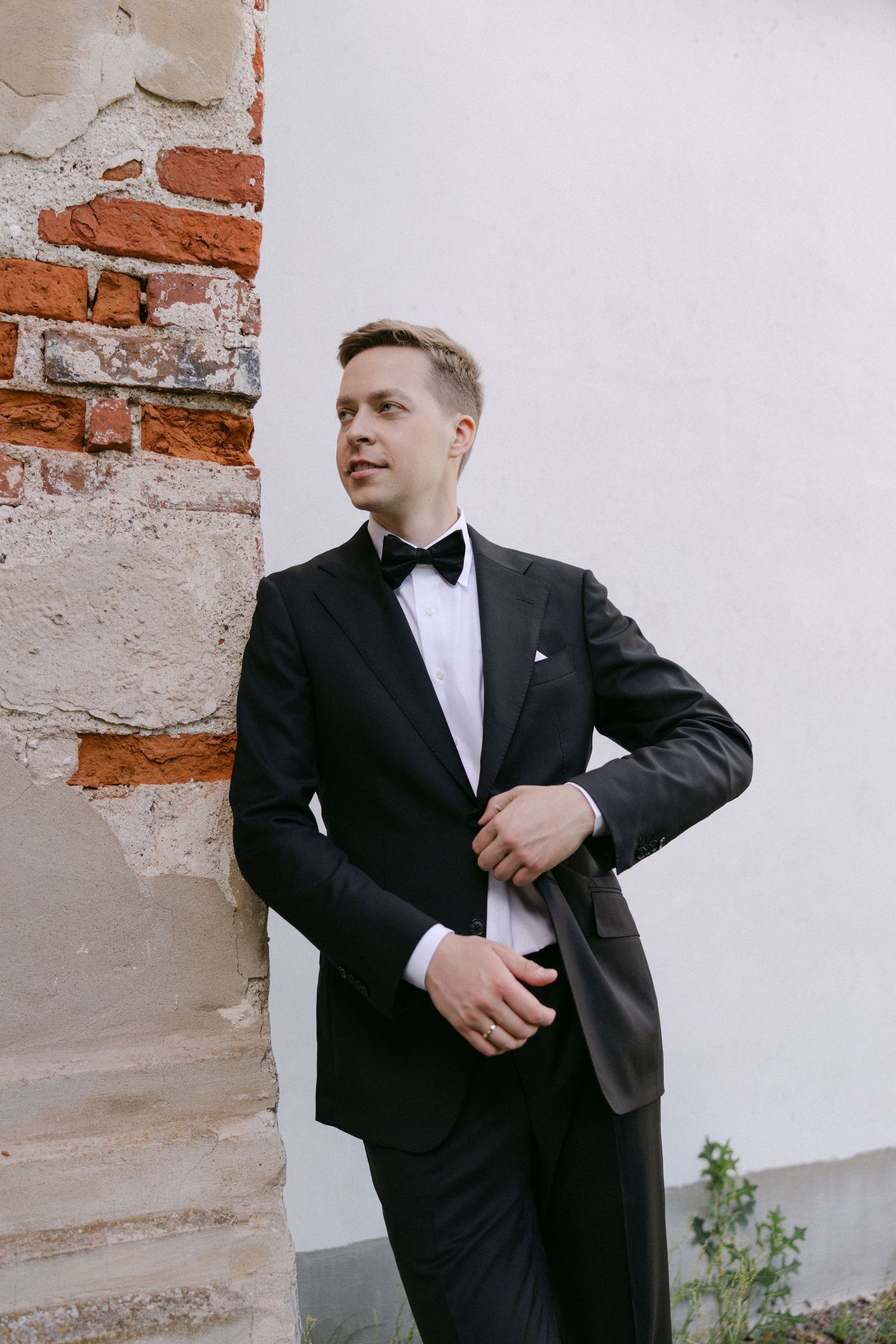 Dapper groom in black tuxedo with bow tie leaning against rustic brick and white wall outdoors.