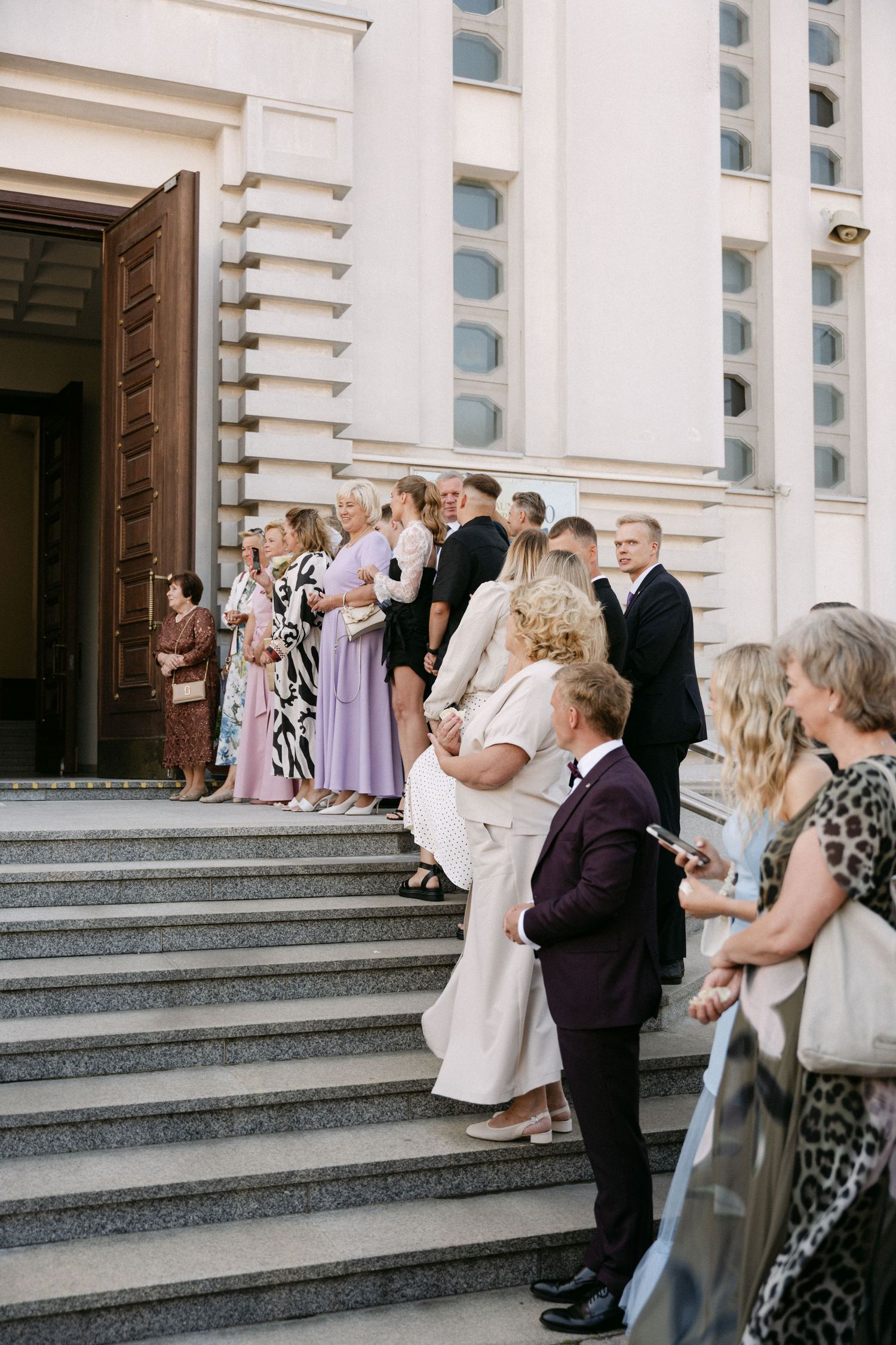Wedding guests in formal attire gathered on stone steps outside an elegant classical building with large wooden doors.