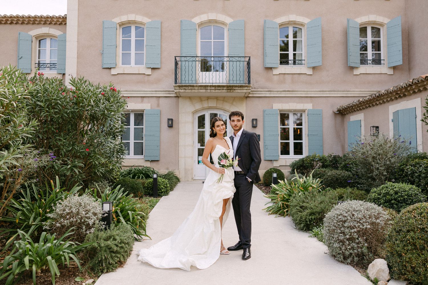 Bride and groom pose together in front of a elegant French villa with blue shutters and lush gardens.