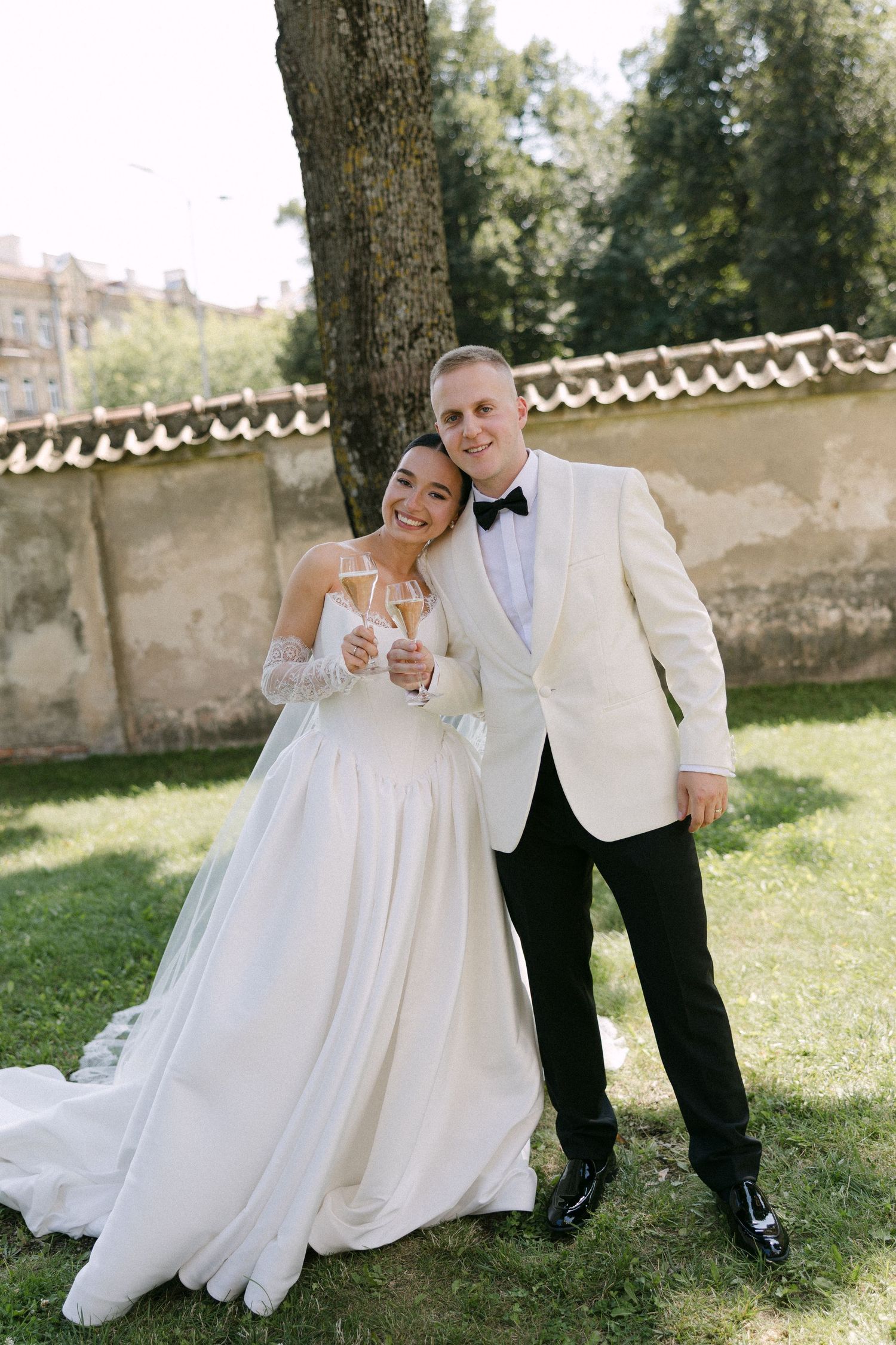 Bride and groom toasting with champagne glasses outdoors near a tree in a lush garden on their wedding day.