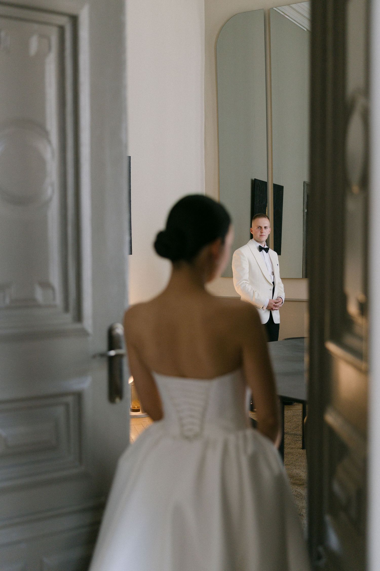 Bride in white strapless gown faces groom in white tuxedo jacket reflected in mirror through elegant doorway.