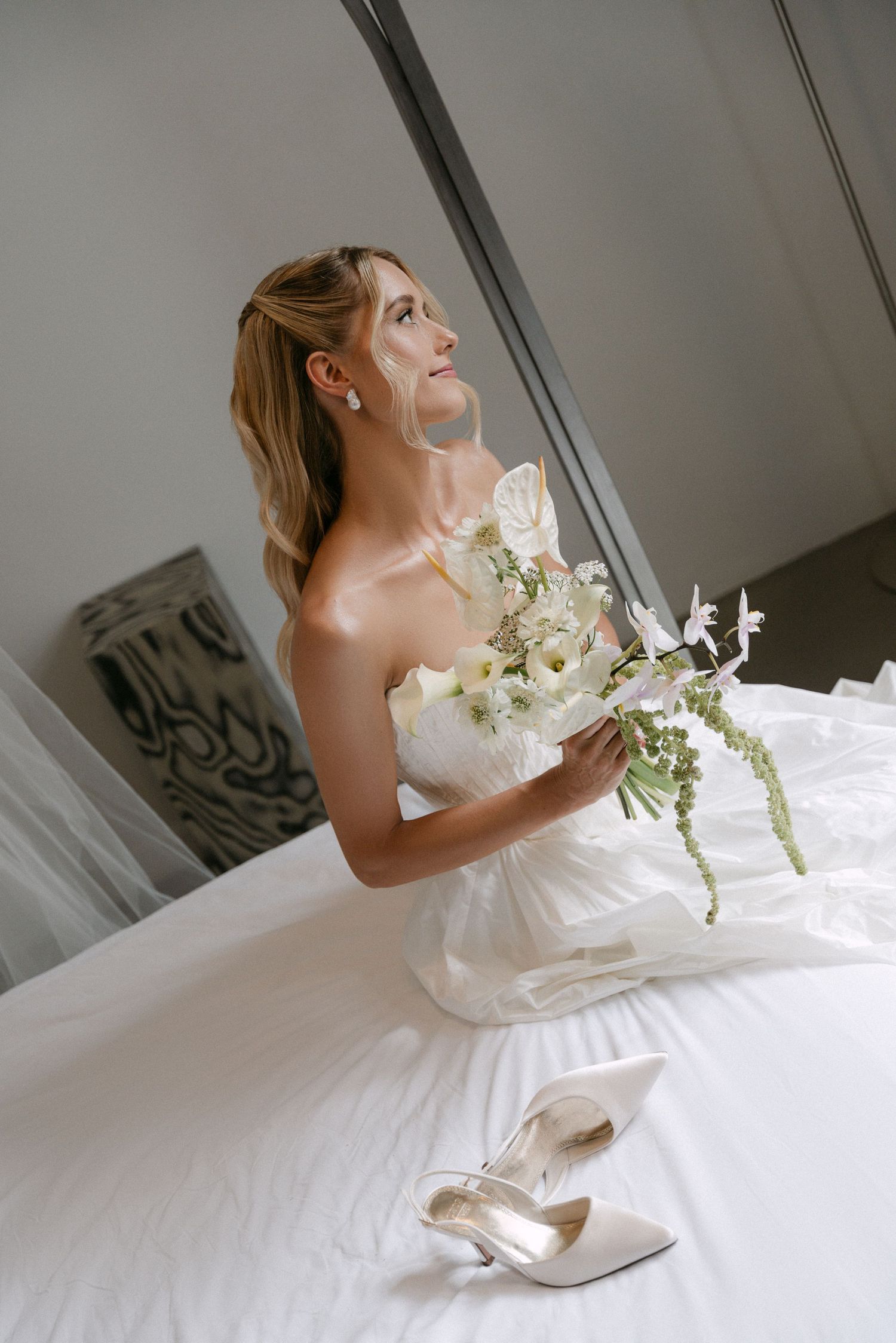 Bride in strapless white gown sitting on bed holding floral bouquet with white heels nearby.