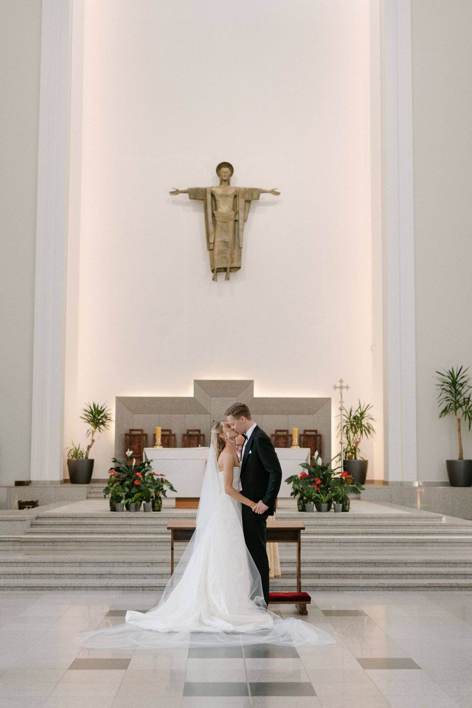 Bride and groom embracing at church altar beneath a Christ statue in a modern white wedding ceremony.