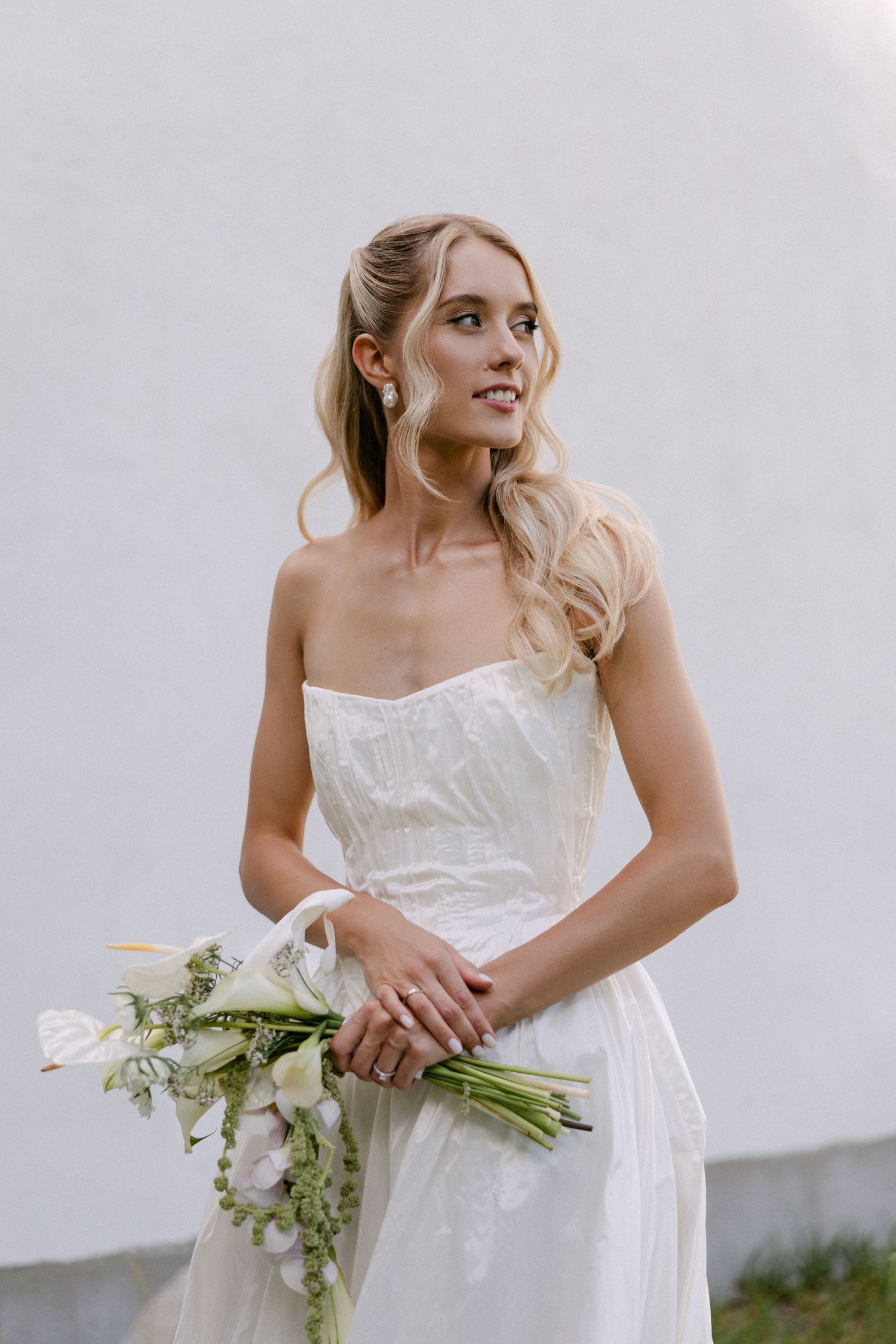 Elegant bride in a strapless white gown holding a white calla lily bouquet, smiling softly against a white wall.