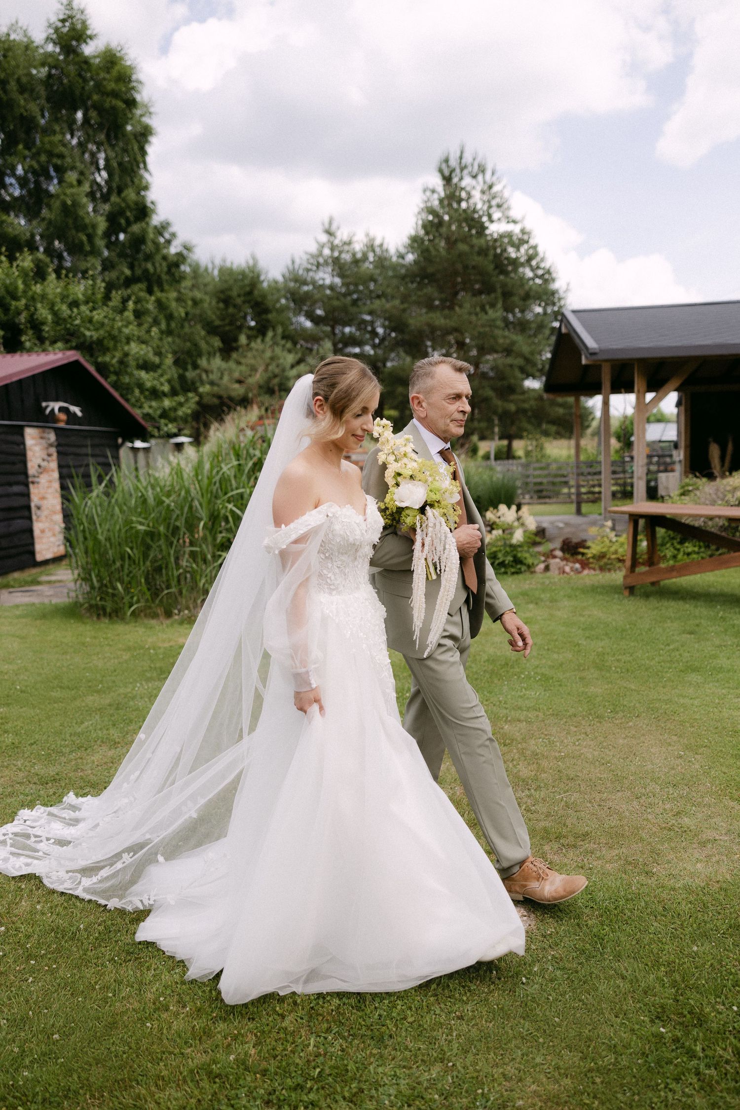 Bride in off-shoulder white gown with long veil walks with escort across green lawn at outdoor wedding.