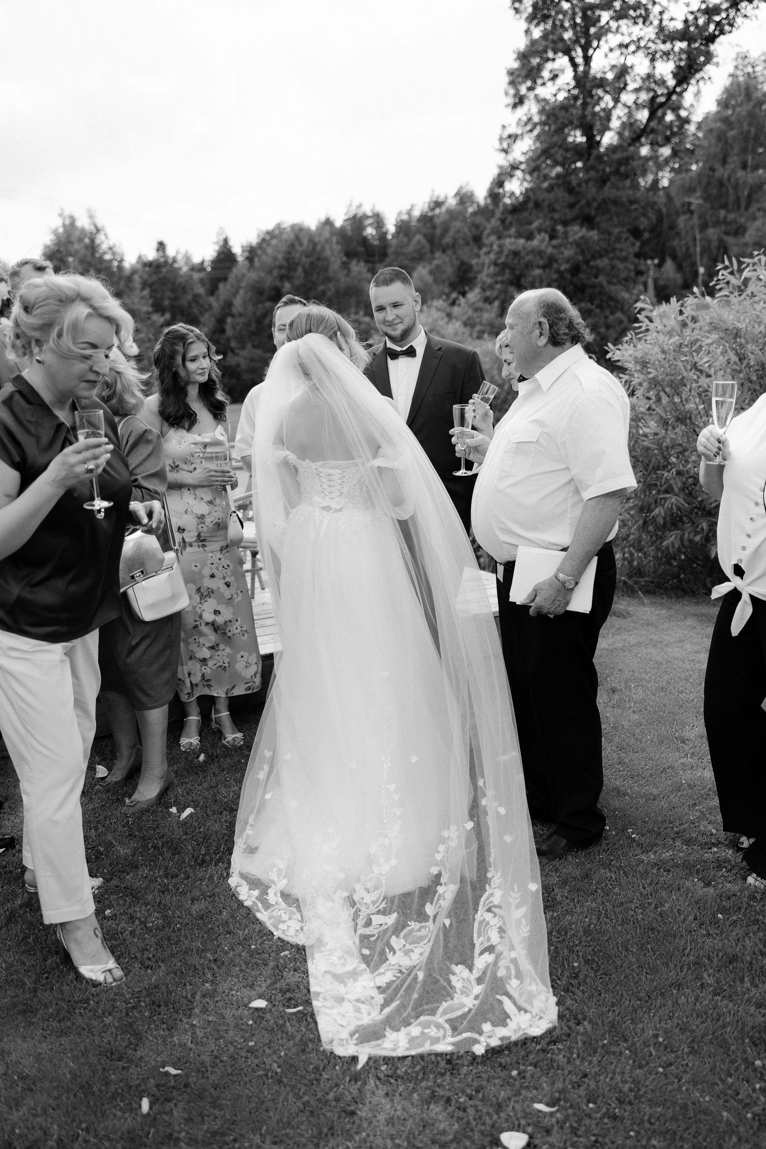 Black and white photo of bride in lace veil chatting with wedding guests outdoors.