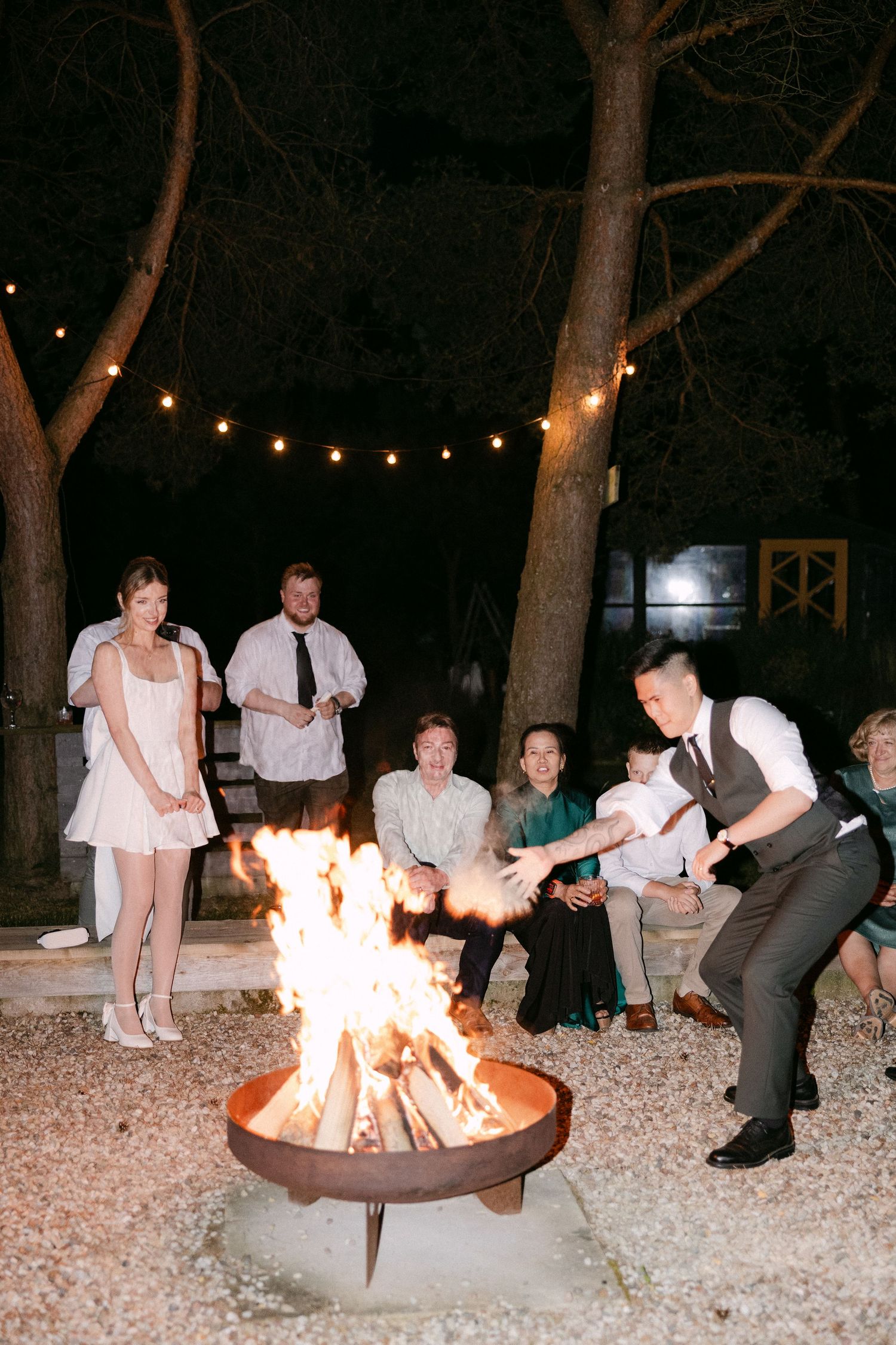 Guests gather around a glowing fire pit at an outdoor wedding reception at night, with string lights illuminating the trees.