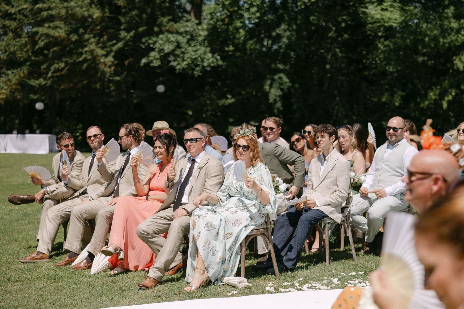 Wedding guests seated outdoors on chairs watching a sunny garden ceremony.