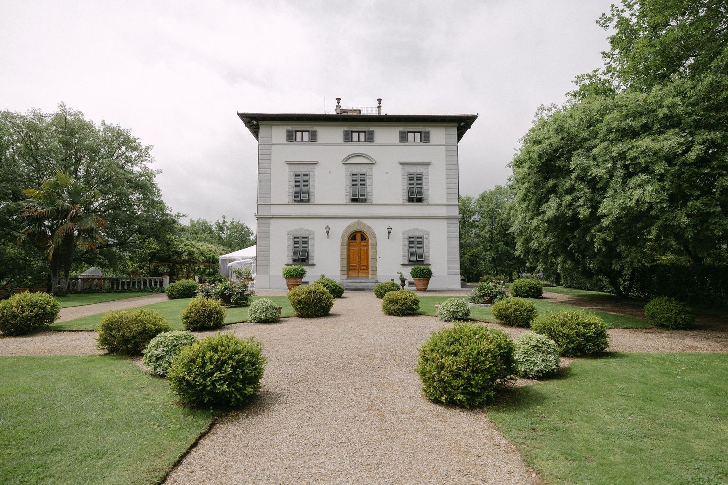 Elegant Italian villa with manicured hedges, gravel circular driveway, and lush green trees under cloudy skies.