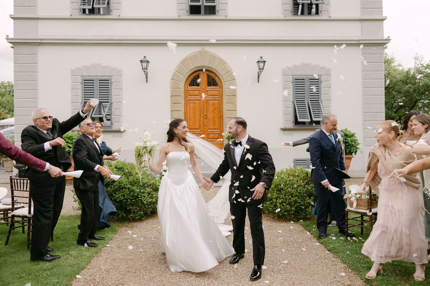 Bride and groom exit ceremony as guests toss white flower petals outside elegant villa.