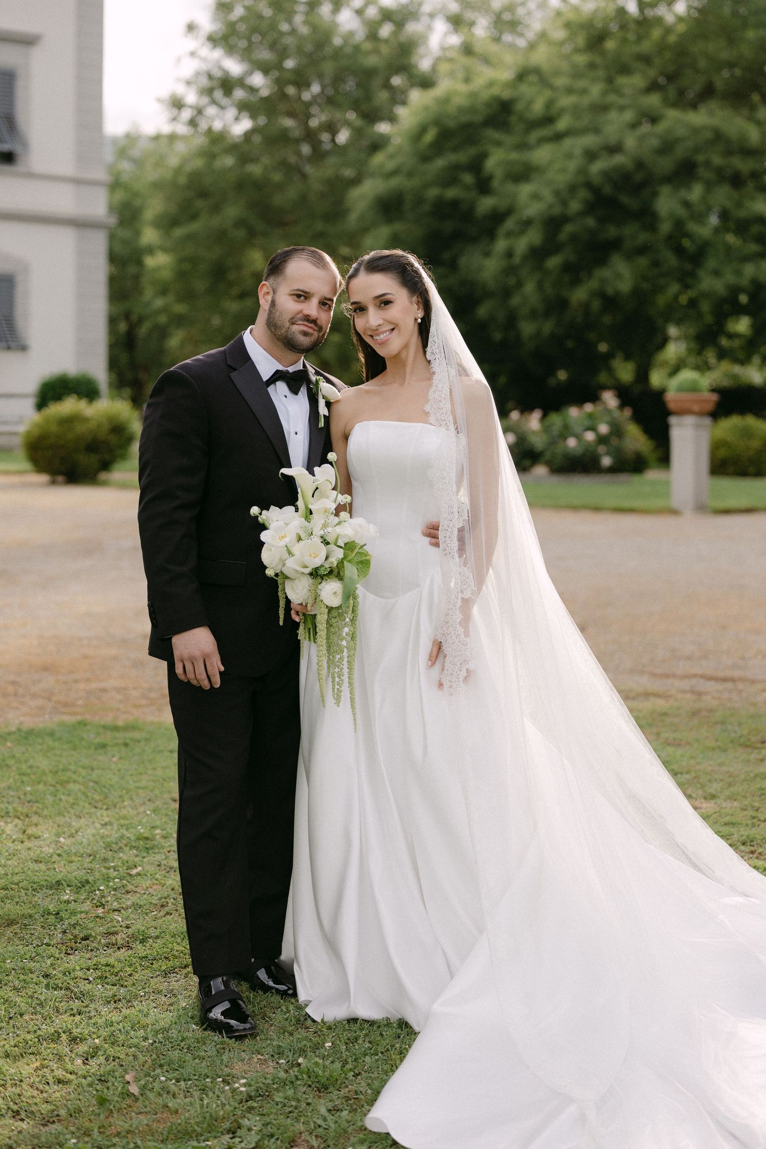 Happy newlywed couple posing together outside elegant Italian villa, bride in flowing white gown with cathedral veil.