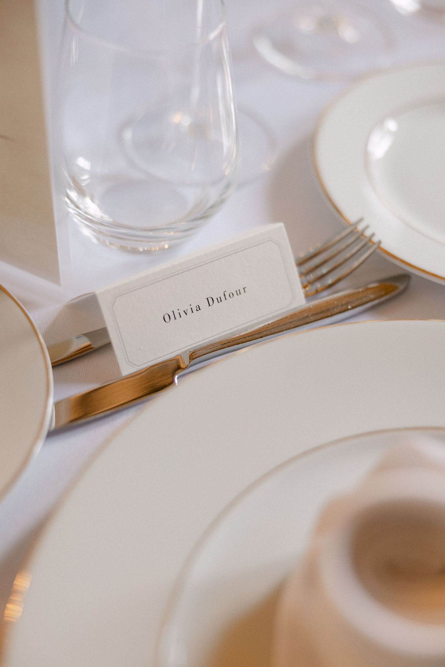 Elegant wedding place card reading Olivia Dufour on white table setting with gold-rimmed plates.