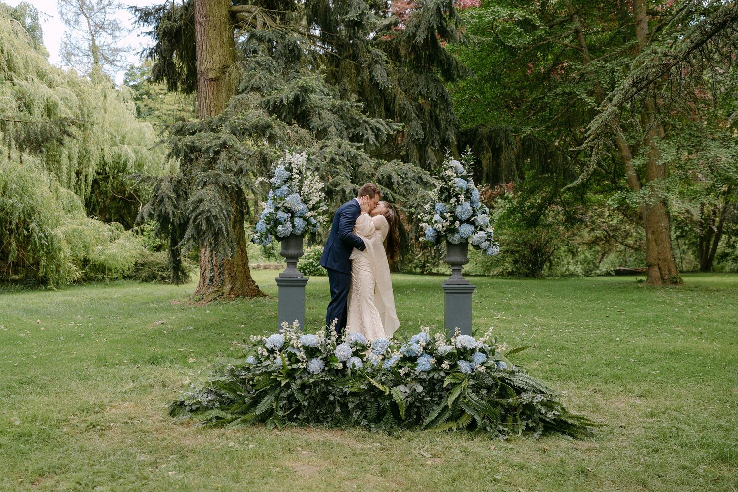 Couple embraces at floral altar with blue hydrangeas in lush garden ceremony.