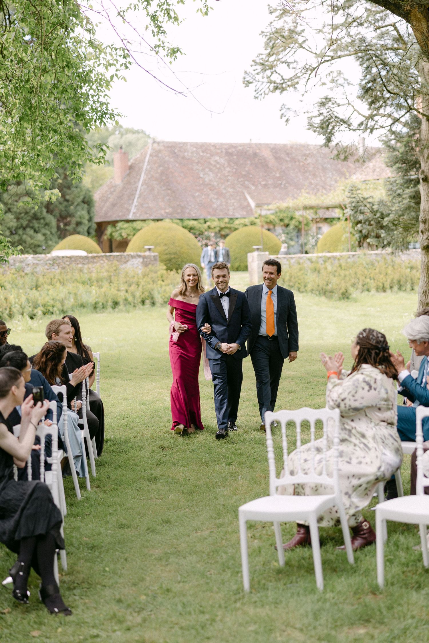 Groom walks down outdoor aisle with two guests as wedding attendees applaud in a lush garden setting.