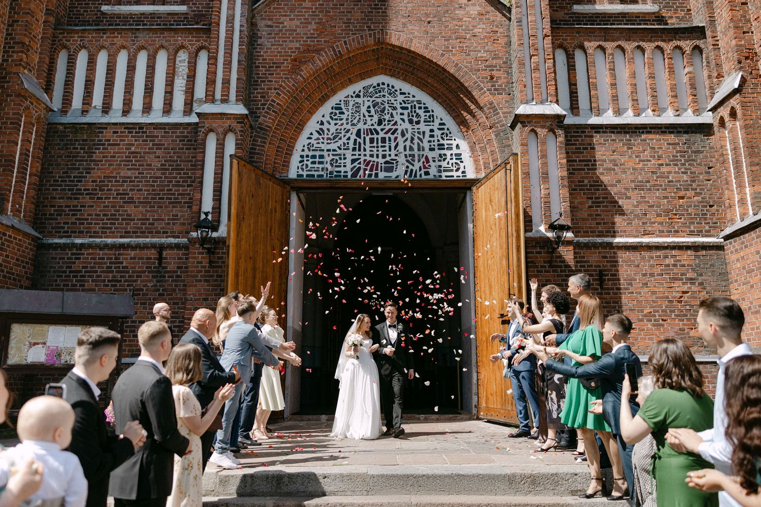 Newlyweds exit a brick church greeted by guests throwing confetti during a sunny outdoor wedding ceremony.
