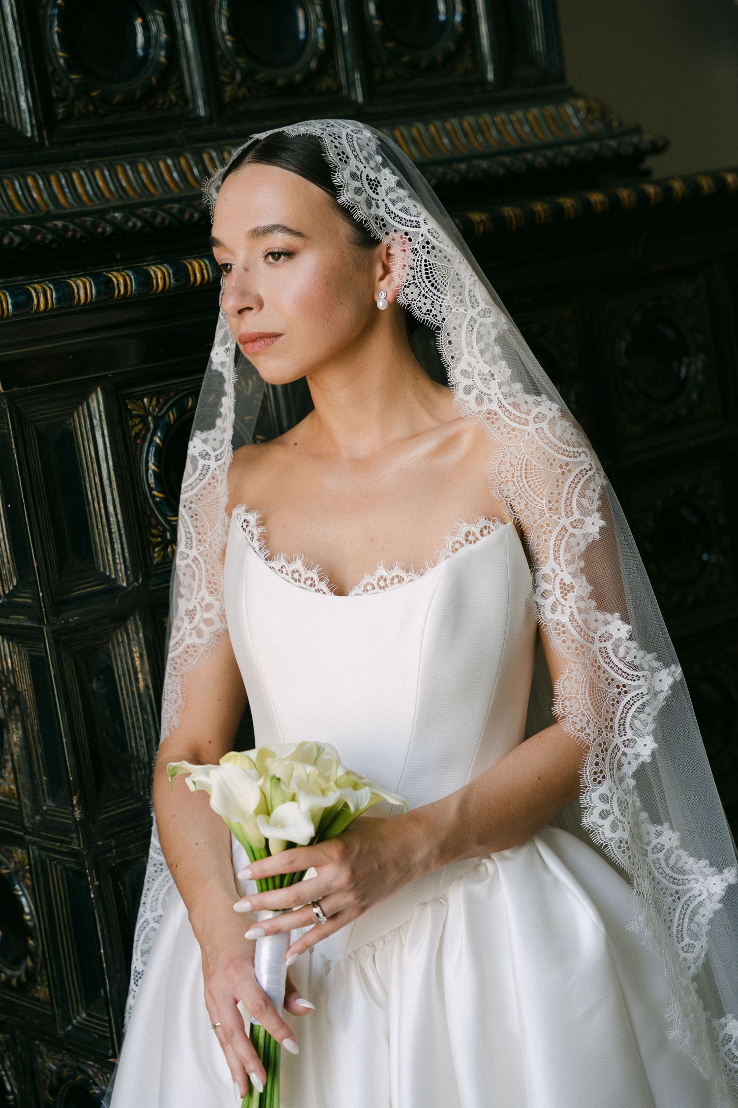 Bride in white strapless gown with lace veil holds calla lily bouquet in elegant interior.