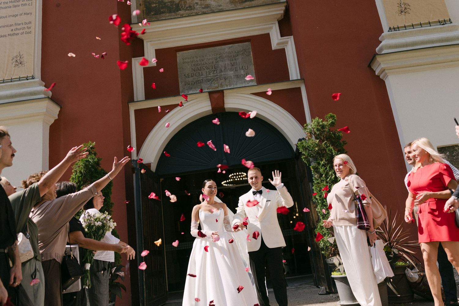 Newlywed couple exits church amid rose petal shower, guests celebrating joyful wedding ceremony outdoors.