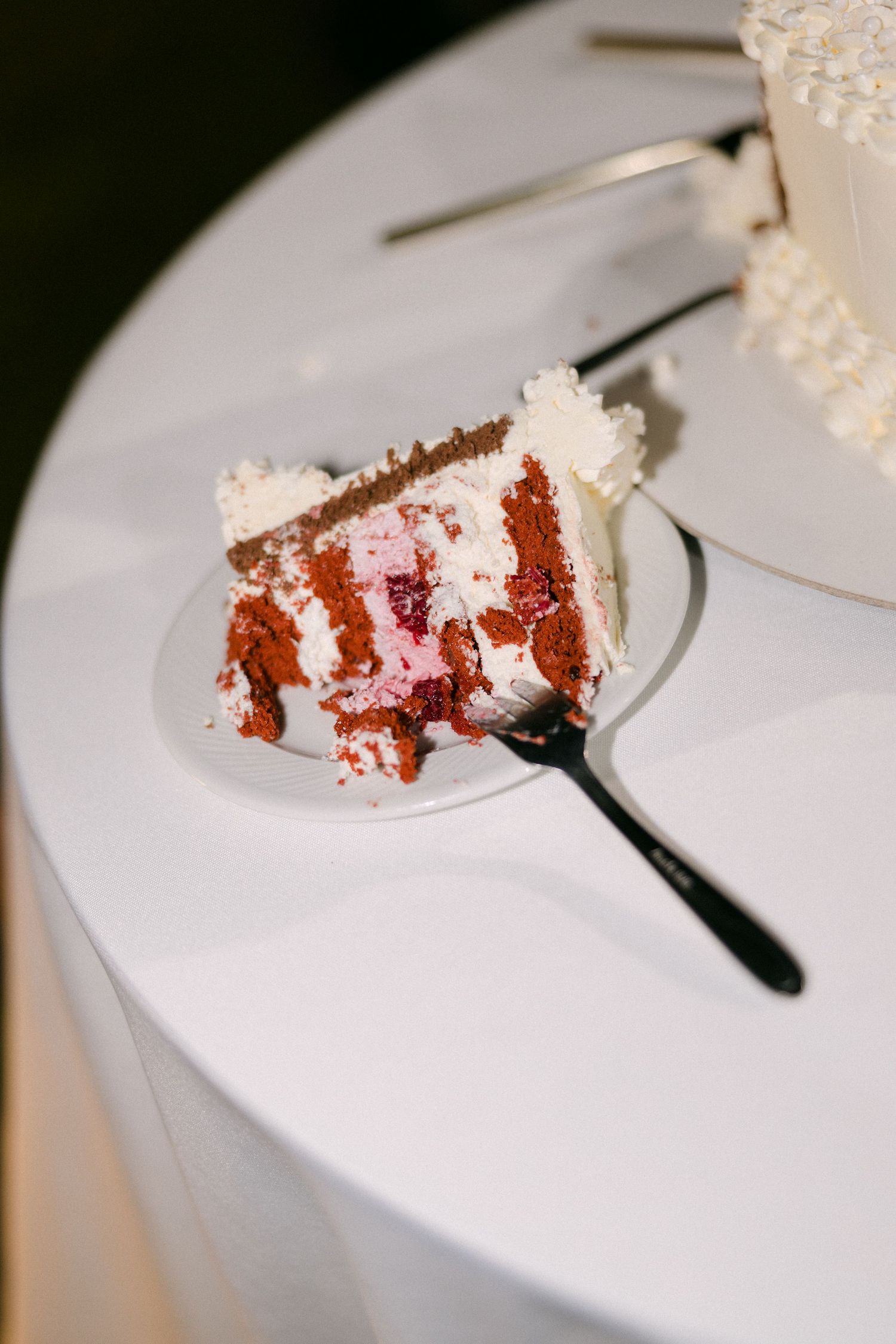 Slice of layered red velvet wedding cake with white frosting on plate with black fork beside tiered cake.