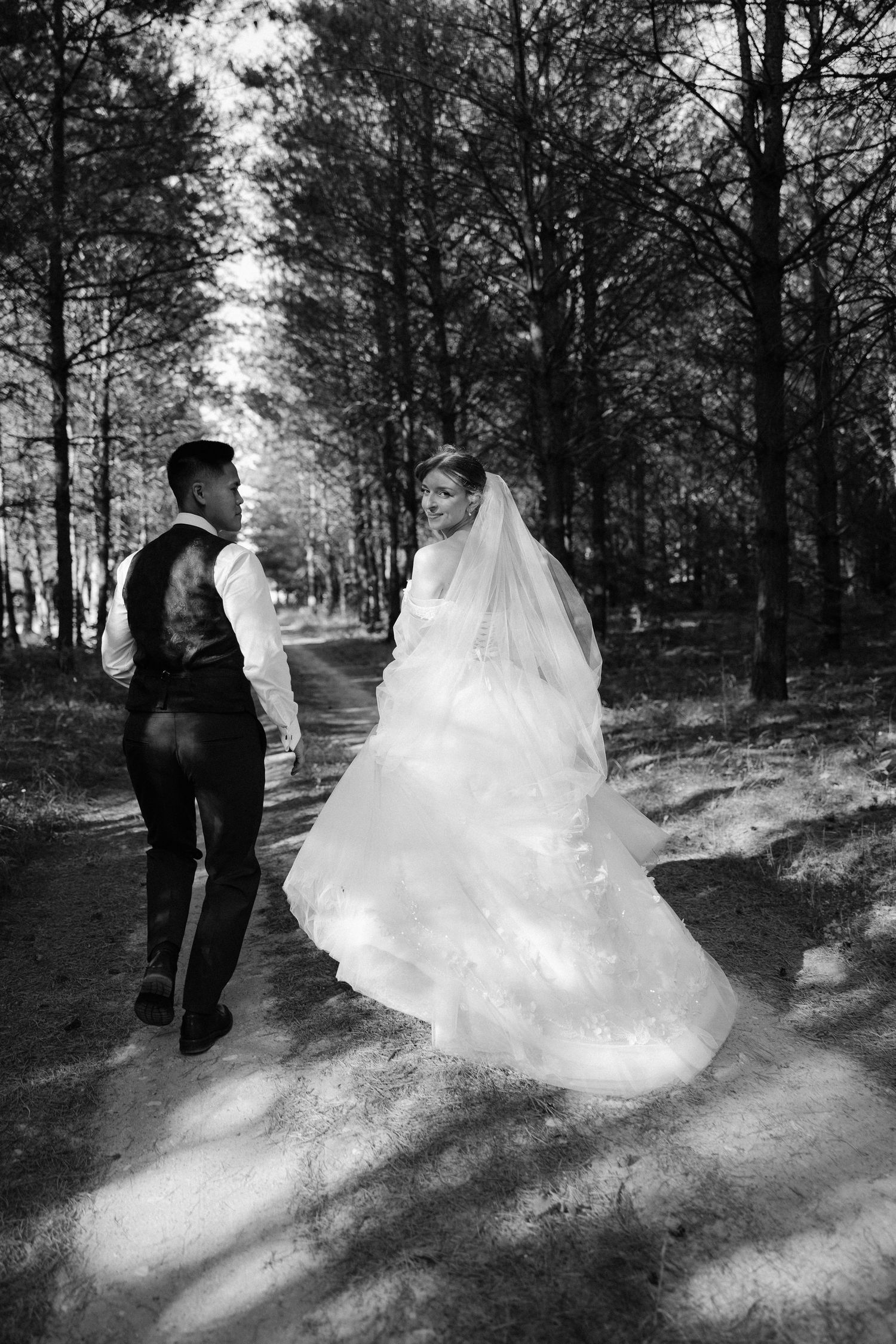 Black and white photo of bride and groom walking along forest path, bride glancing back at camera.