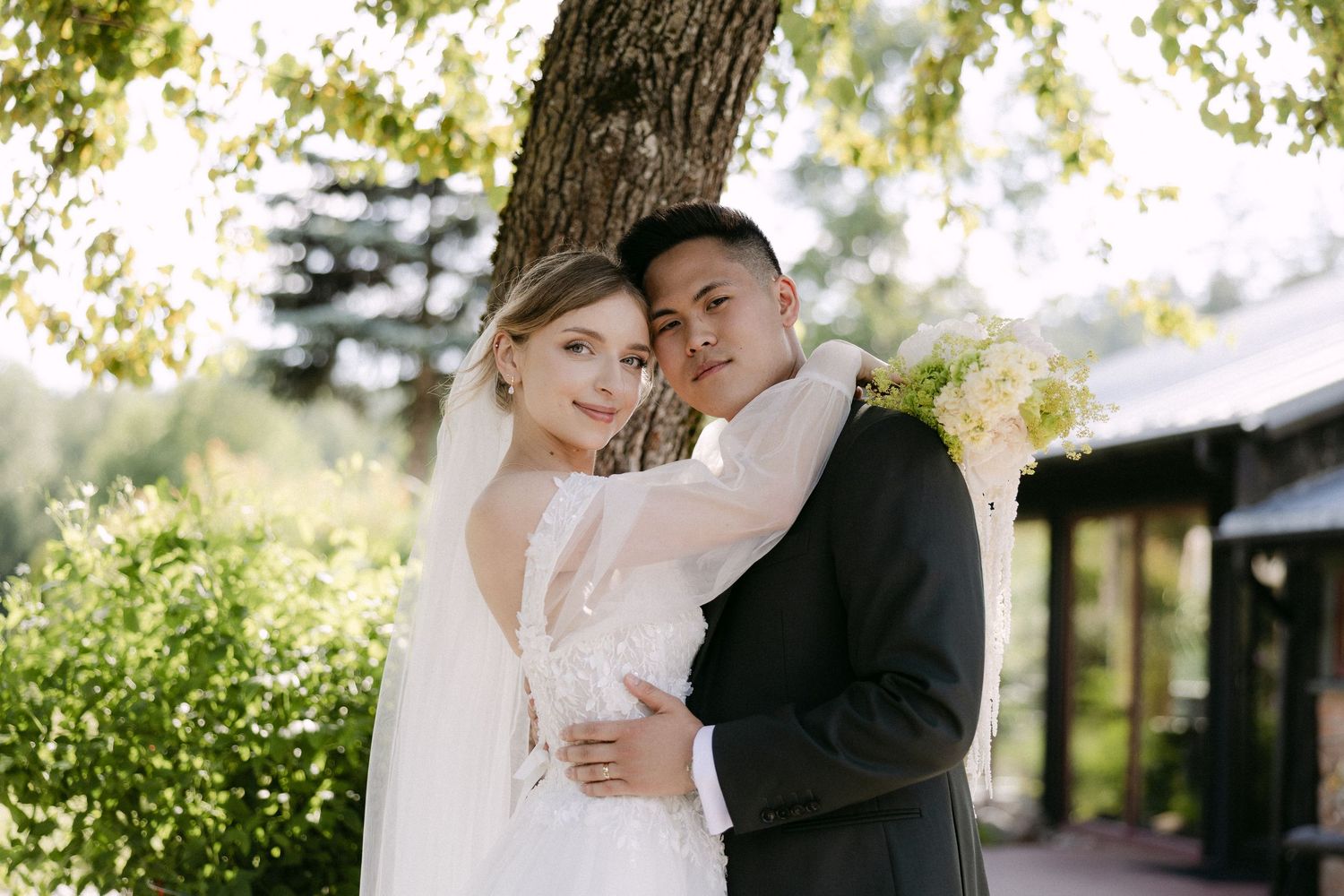 Bride and groom embracing under a tree in golden sunlight, bride wearing a lace gown and veil.