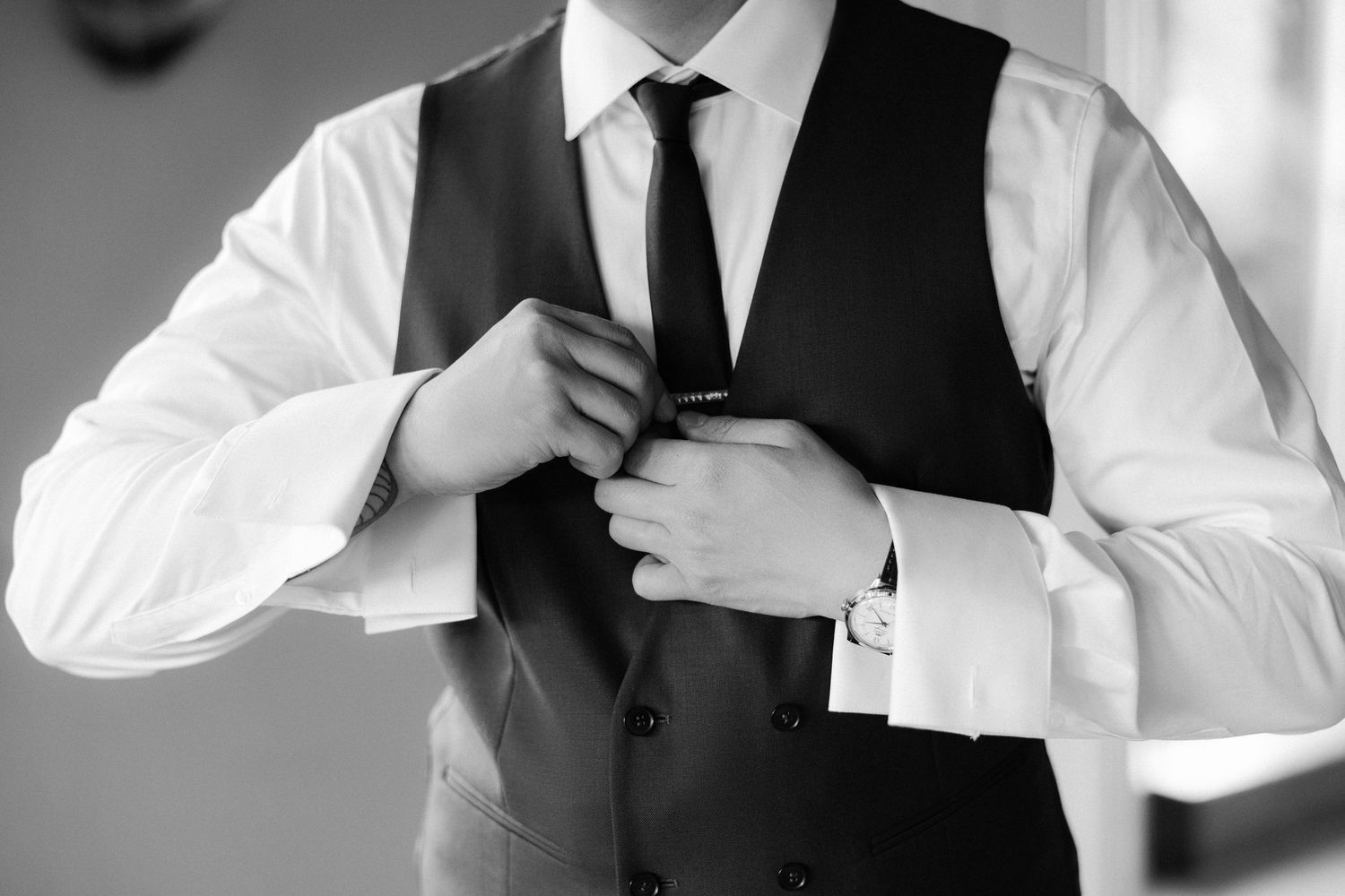 Black and white photo of a groom adjusting his tie while wearing a vest, dress shirt, and cufflinks.