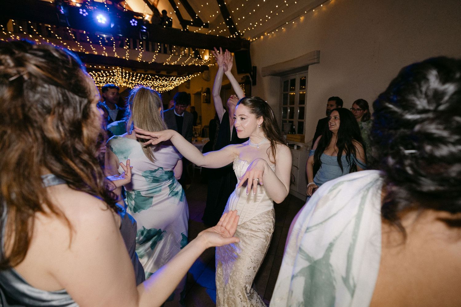 Bride in sequin gown dancing joyfully with guests under fairy lights at wedding reception.