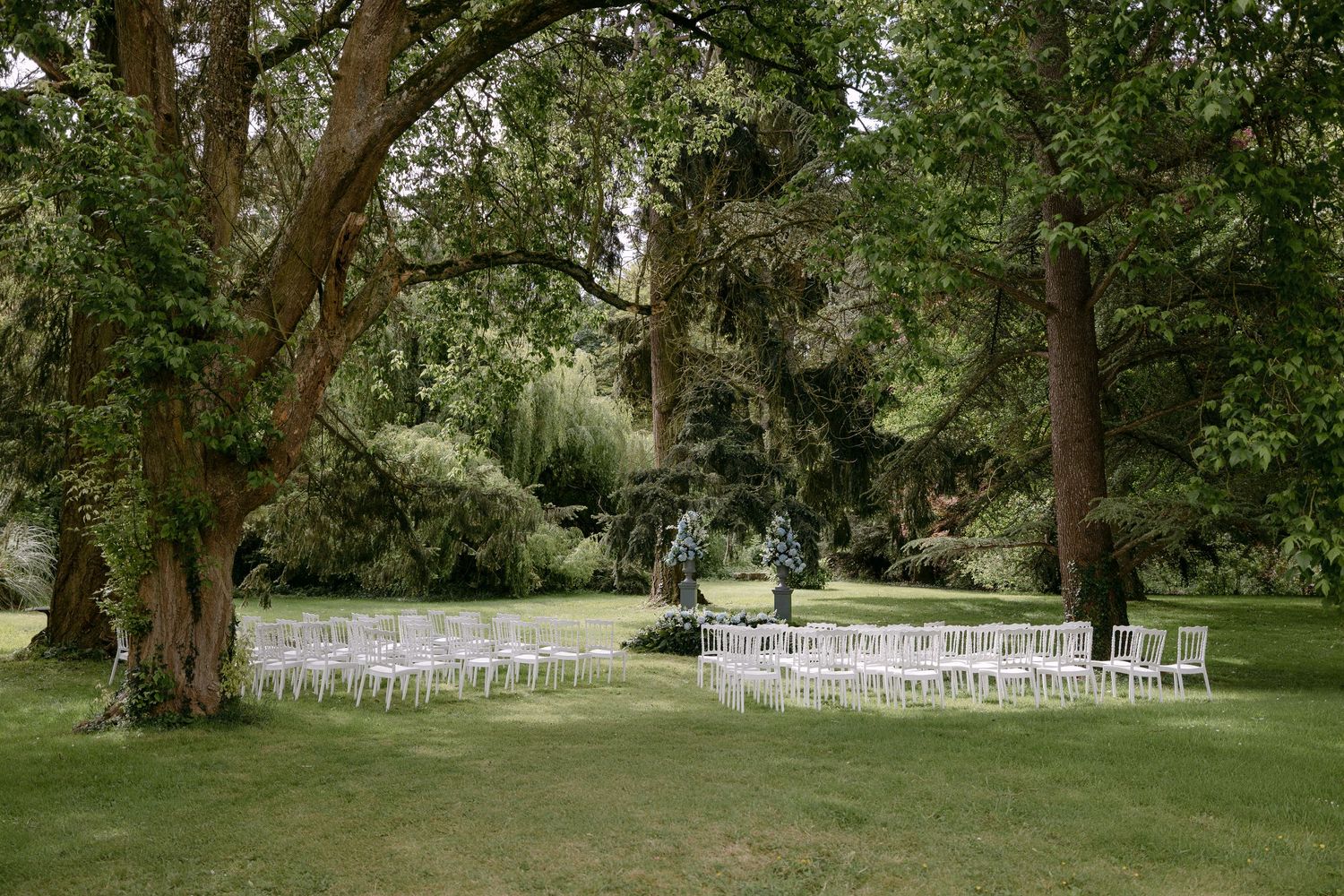Outdoor wedding ceremony setup with white chairs arranged on lush green lawn surrounded by tall trees.