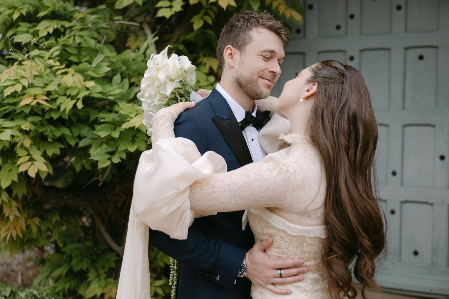 Bride and groom sharing a romantic embrace outdoors, bride holding white hydrangea bouquet in lace gown.