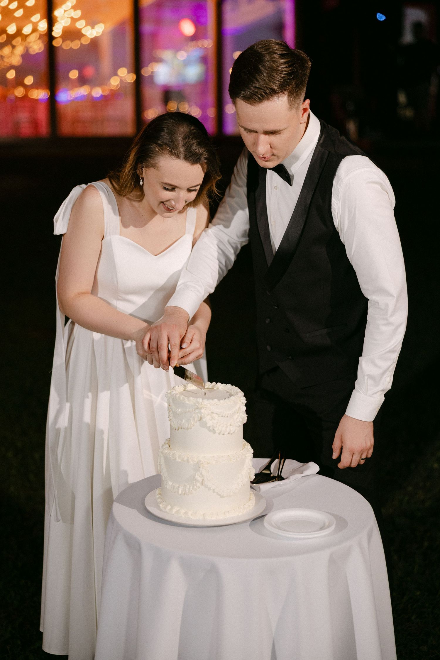 Bride and groom cutting elegant two-tier white wedding cake at night reception.