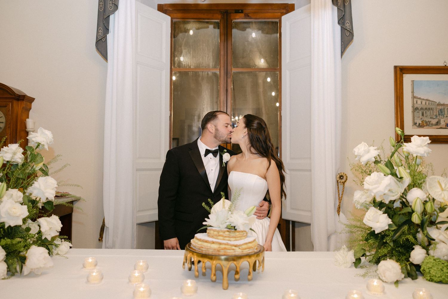 Newlyweds sharing romantic kiss over gold drip wedding cake surrounded by white floral arrangements and candles.