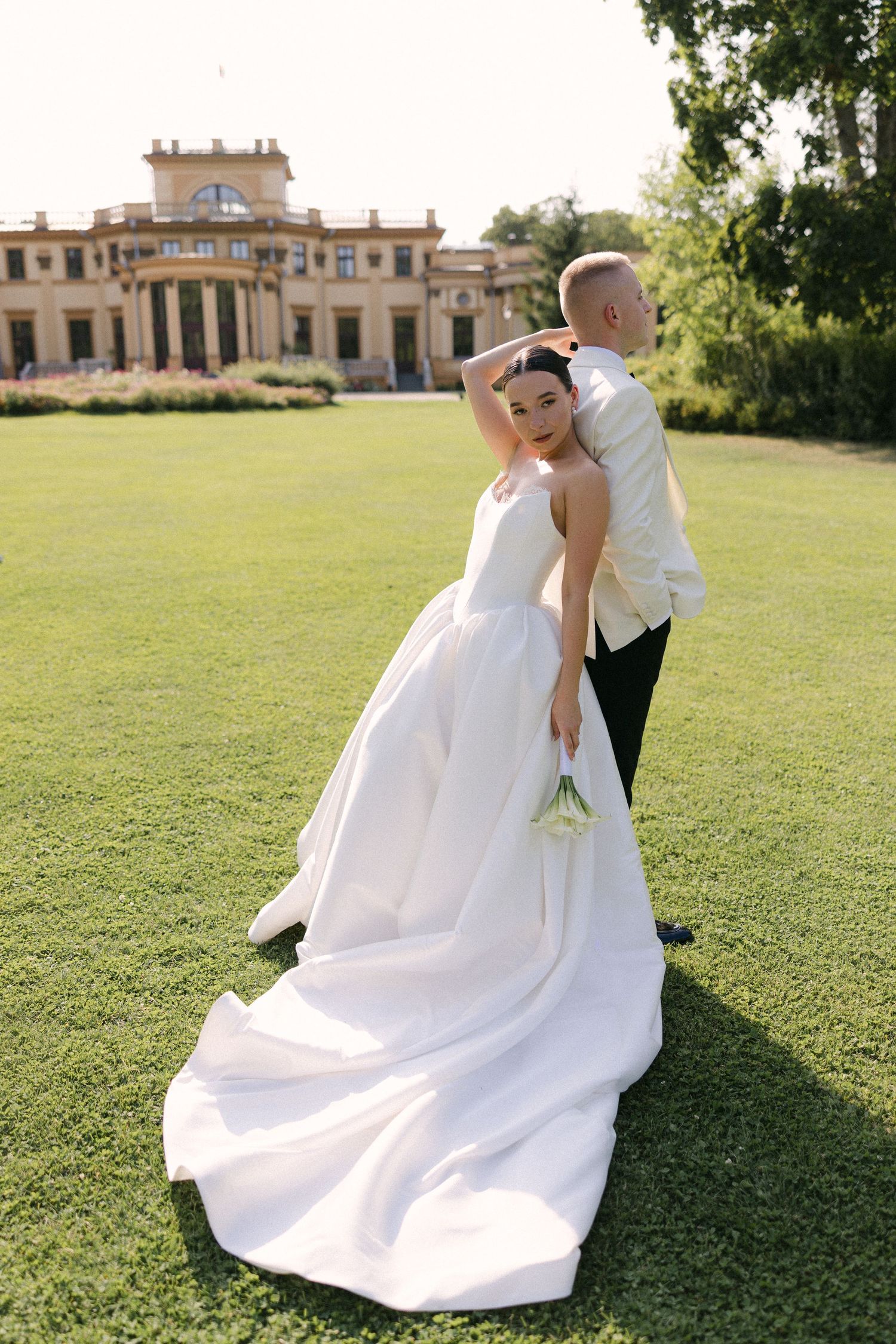 Elegant bride in white ball gown looking back at camera while groom embraces her before luxury manor.