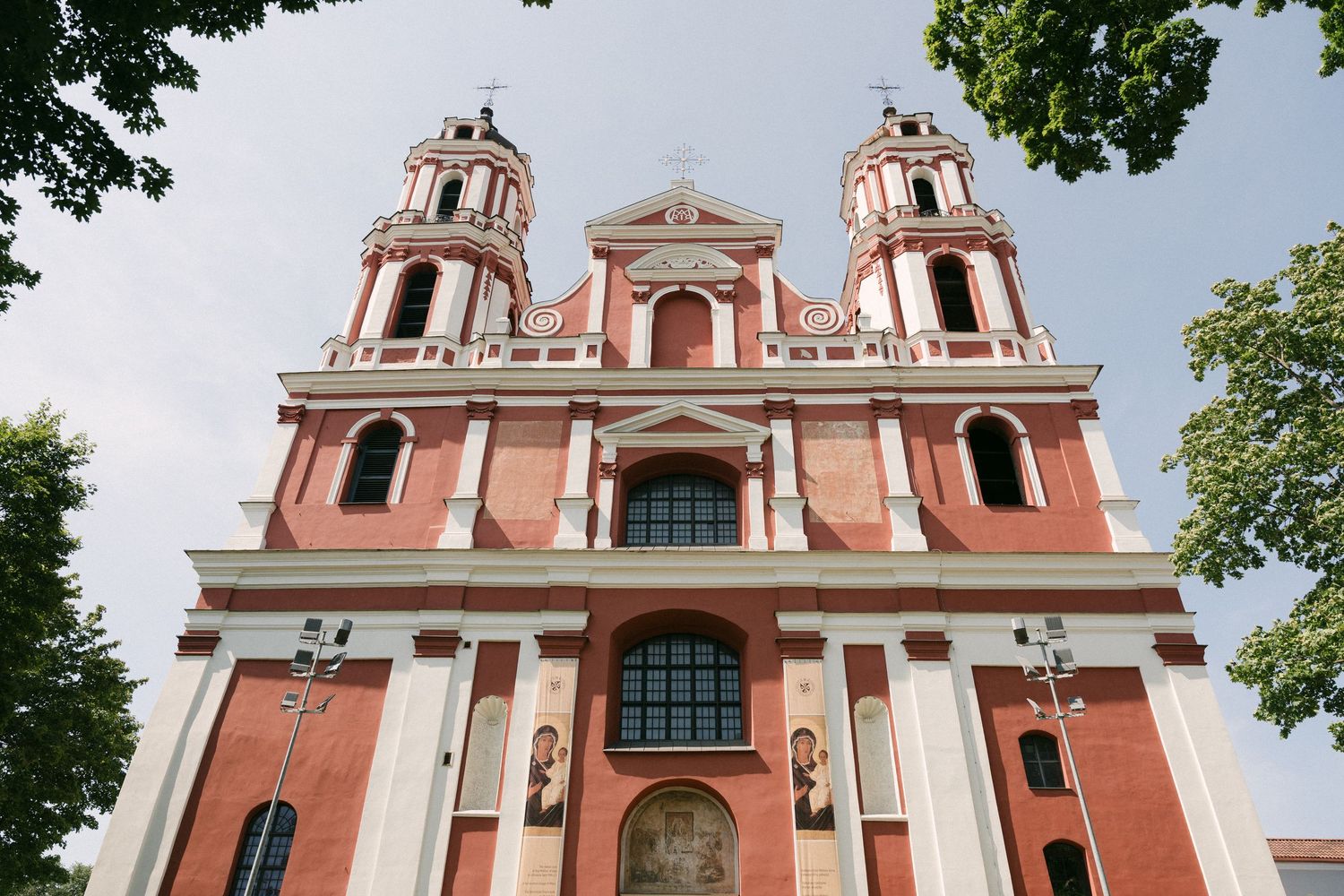 Red Baroque church with twin bell towers against a cloudy sky, surrounded by green trees in Vilnius, Lithuania.