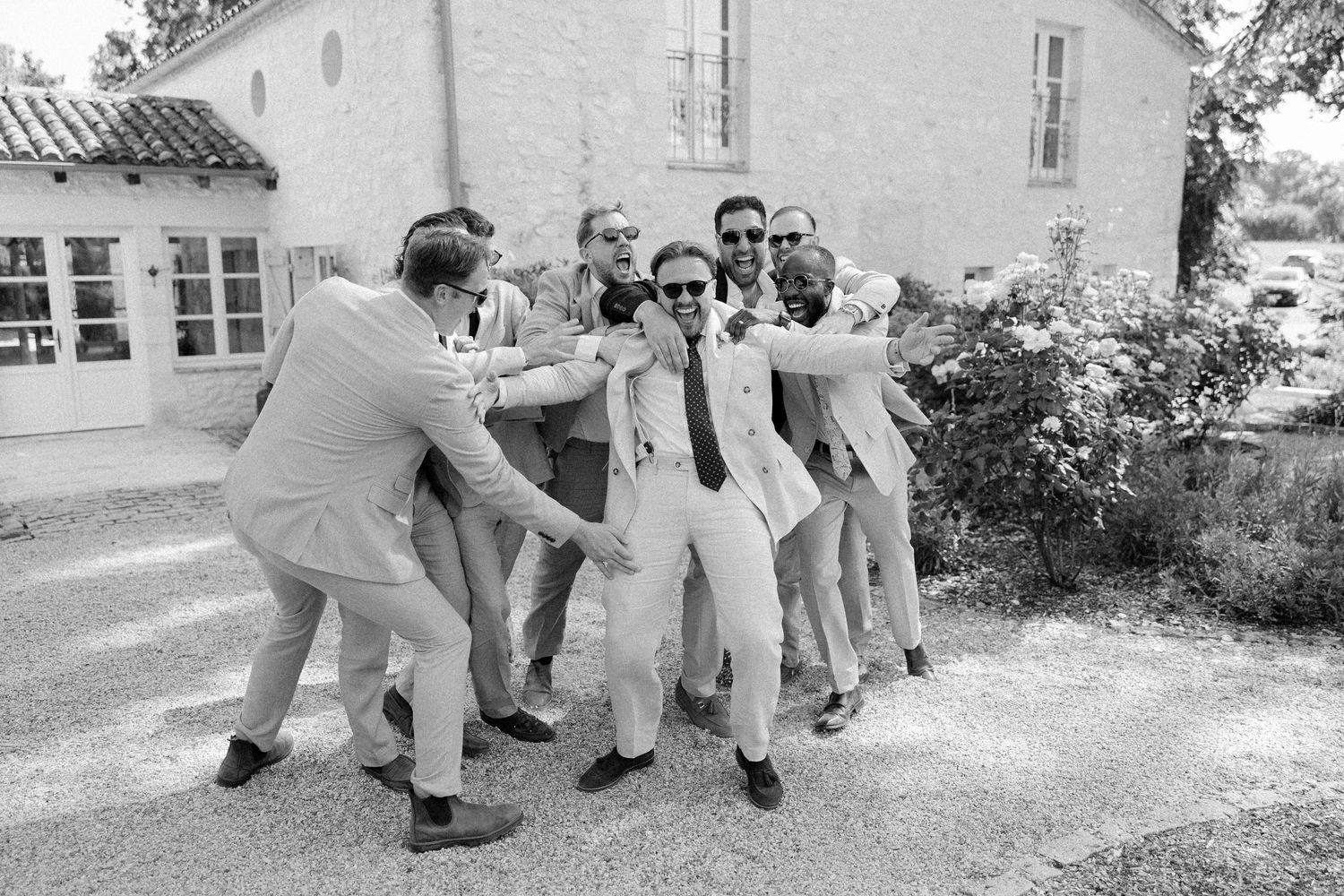 Black and white photo of groomsmen playfully surrounding groom outside wedding venue.